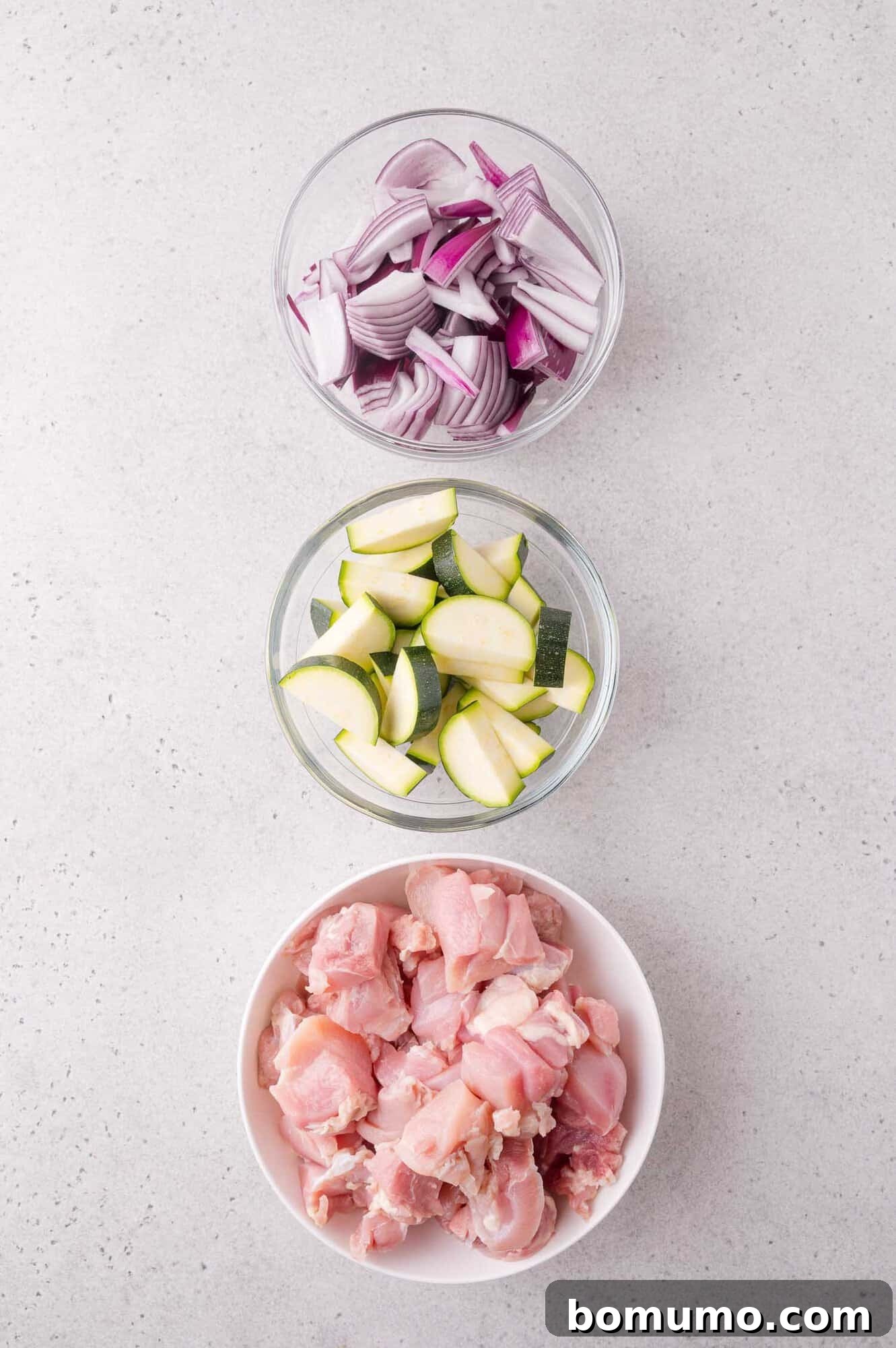 Three bowls containing chopped red onions, sliced zucchini, and raw diced chicken arranged vertically on a light gray surface.