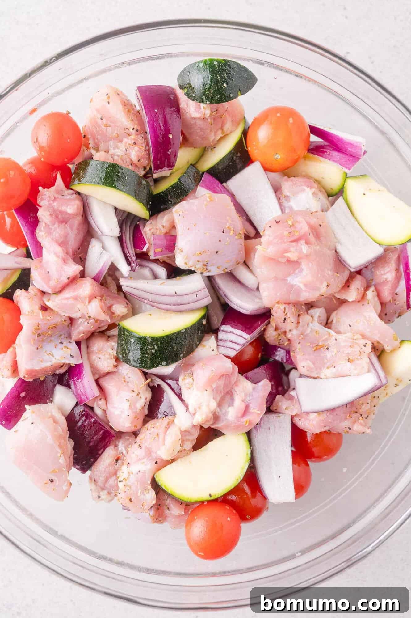 A glass bowl filled with raw chicken pieces, cherry tomatoes, zucchini slices, and red onion chunks, all seasoned and ready for cooking.
