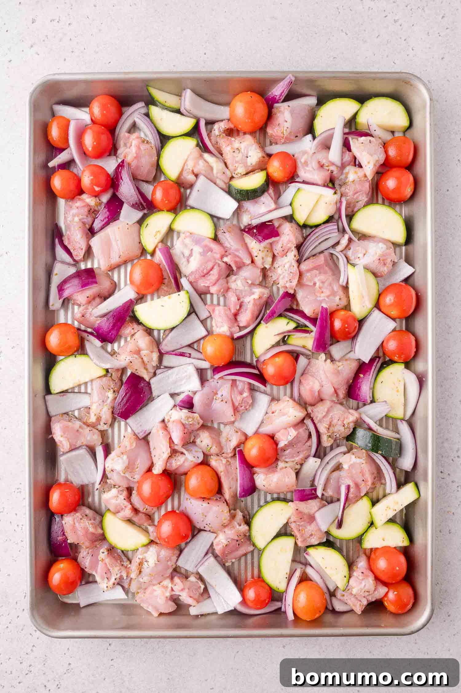 Raw chicken pieces, cherry tomatoes, red onion slices, and zucchini chunks arranged on a metal baking sheet, ready for roasting.