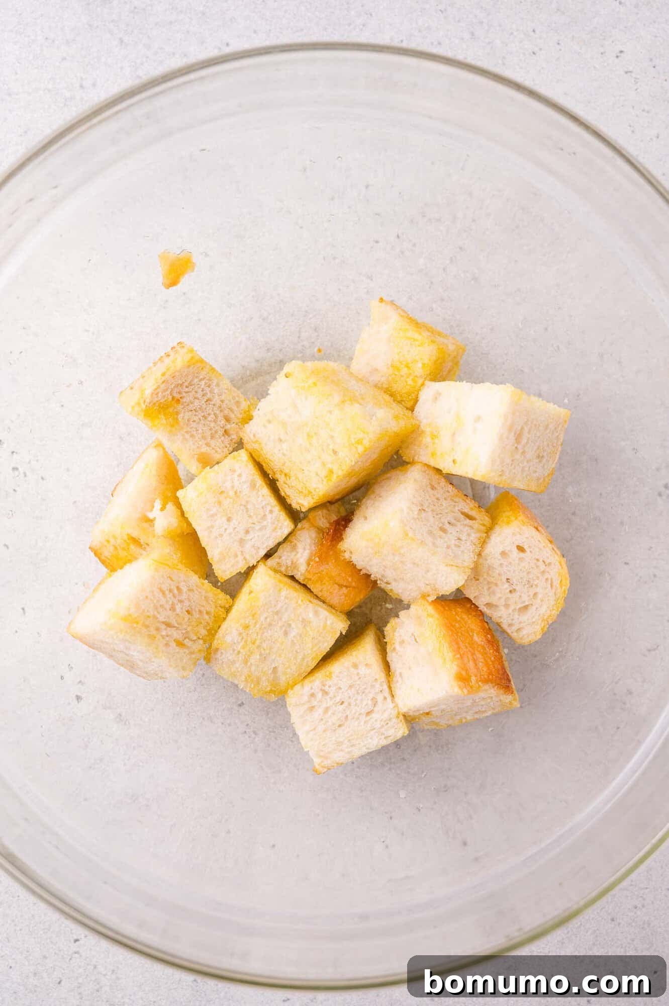 Cubed pieces of bread coated with oil or butter in a clear glass mixing bowl on a light-colored surface.