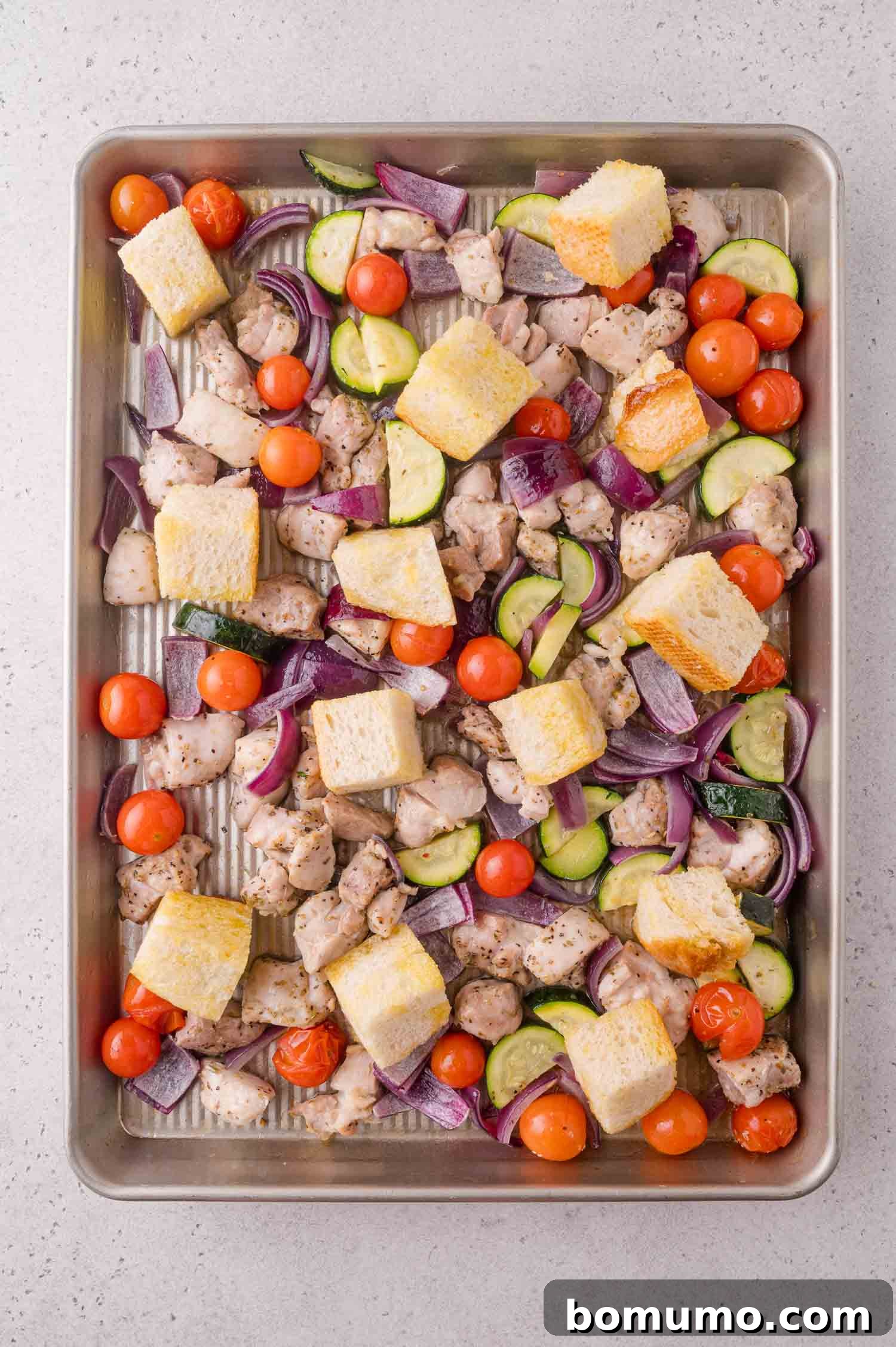 Sheet pan with diced chicken, cherry tomatoes, zucchini slices, red onion pieces, and cubes of bread, all seasoned and ready to be roasted.