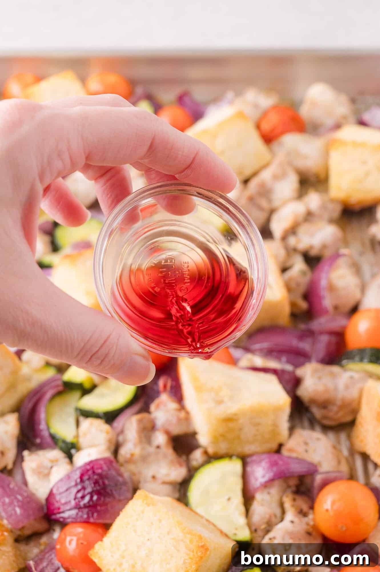 A hand pours red wine vinegar from a small glass bowl over a sheet pan filled with chopped vegetables, bread cubes, and pieces of chicken.