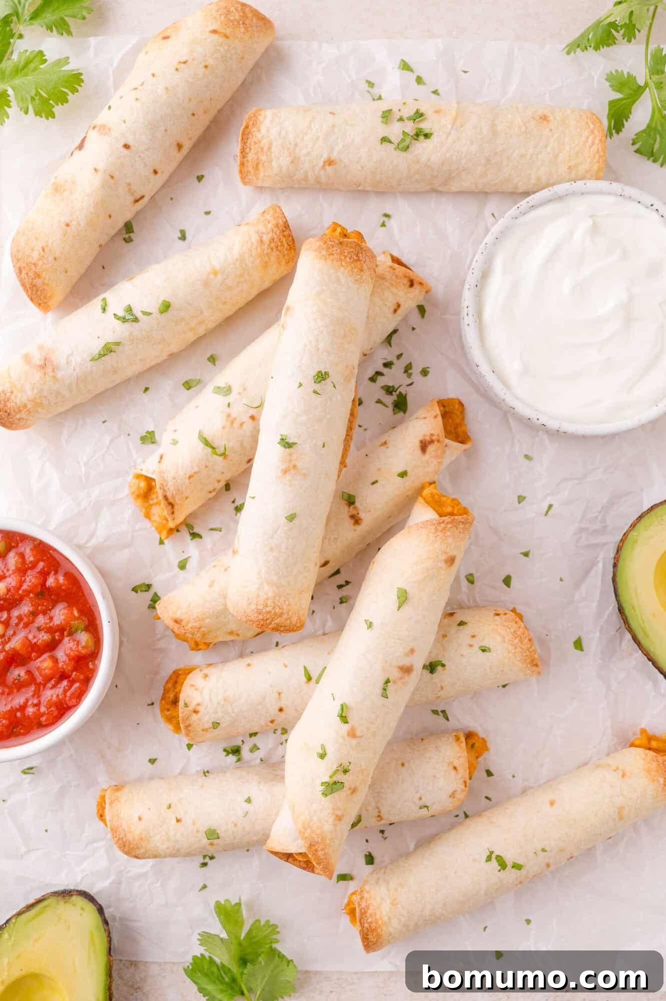 A pile of baked taquitos on parchment paper, garnished with chopped herbs, served with bowls of salsa and sour cream, and avocado halves.