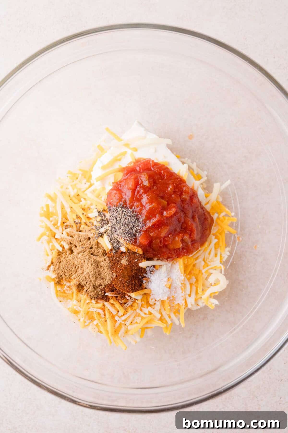Clear glass bowl with shredded cheese, salsa, mayonnaise, and various spices on a light surface, ready to be mixed.