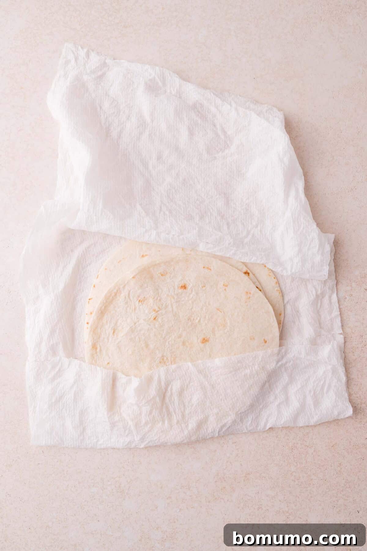 Two flour tortillas partially wrapped in a white paper towel, placed on a light-colored surface.