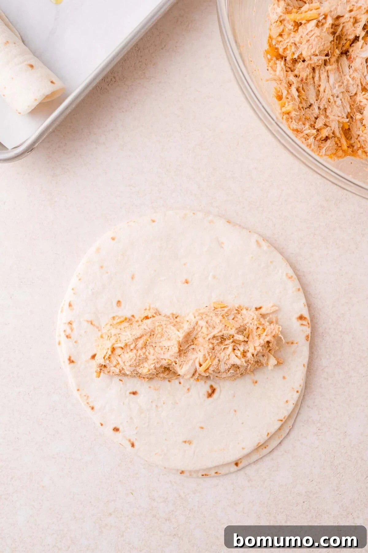 Flour tortilla with a portion of shredded chicken filling in the center, next to a bowl of chicken mixture and a baking sheet with a rolled tortilla.