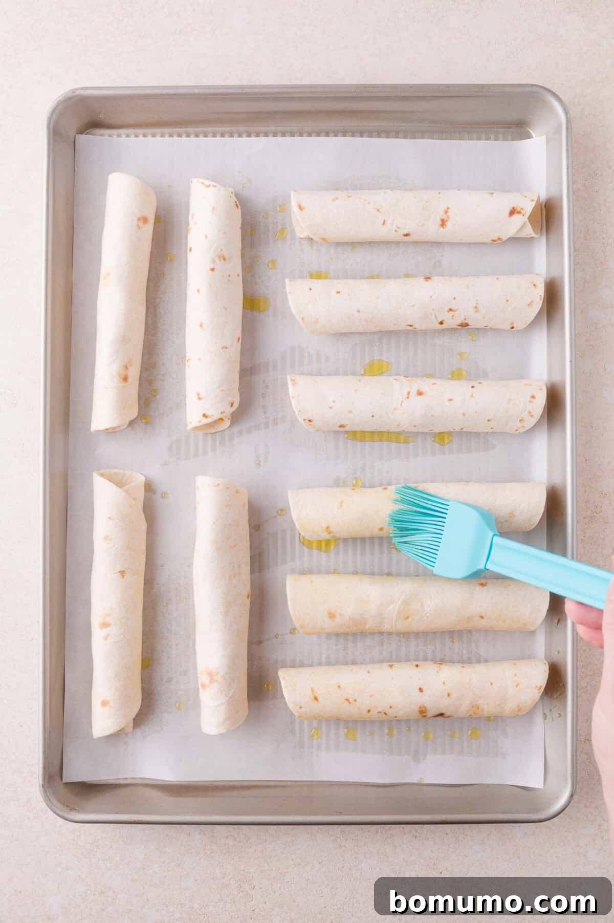 A hand brushes oil onto rolled tortillas arranged on a parchment-lined baking sheet.