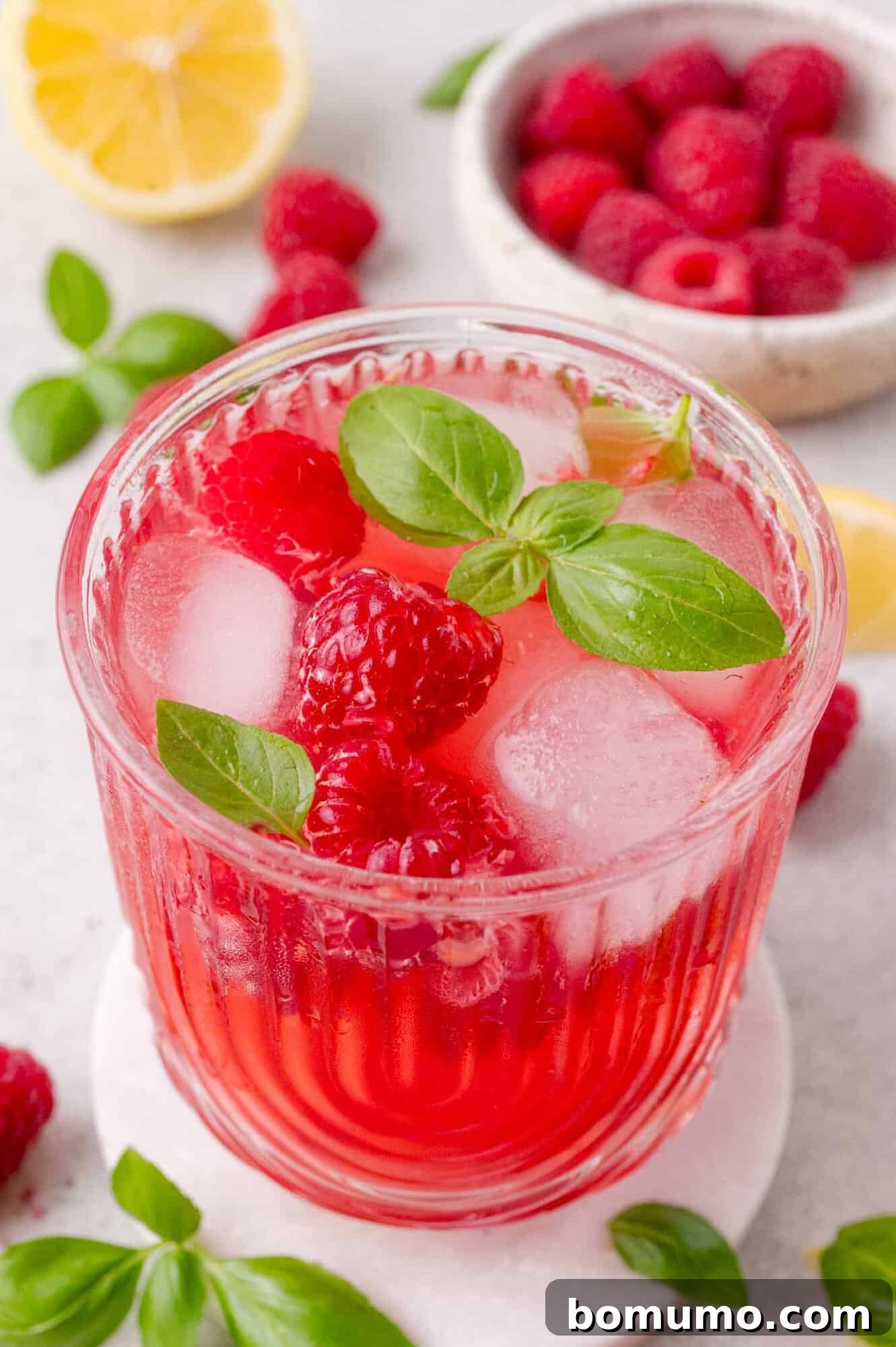 A glass of dark pink raspberry smash mocktail with ice cubes, fresh raspberries, and basil leaves, placed on a white coaster with raspberries and a lemon half in the background.