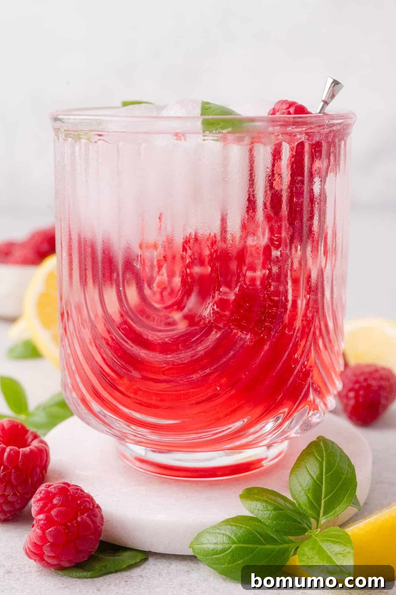 A glass of iced raspberry mocktail garnished with fresh raspberries, basil leaves, and a metal straw, surrounded by lemon slices and basil on a white surface.