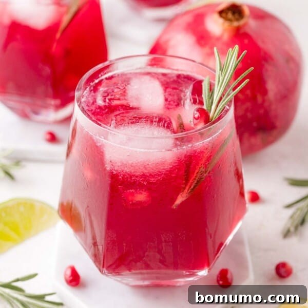 A glass of red pomegranate spritzer with ice and a rosemary sprig garnish sits on a coaster, with a whole pomegranate and lime slices in the background.
