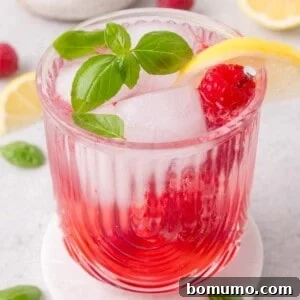 Glass of raspberry beverage with ice, a lemon slice, raspberry, and fresh basil on top, placed on a white coaster with lemon slices and basil leaves in the background.