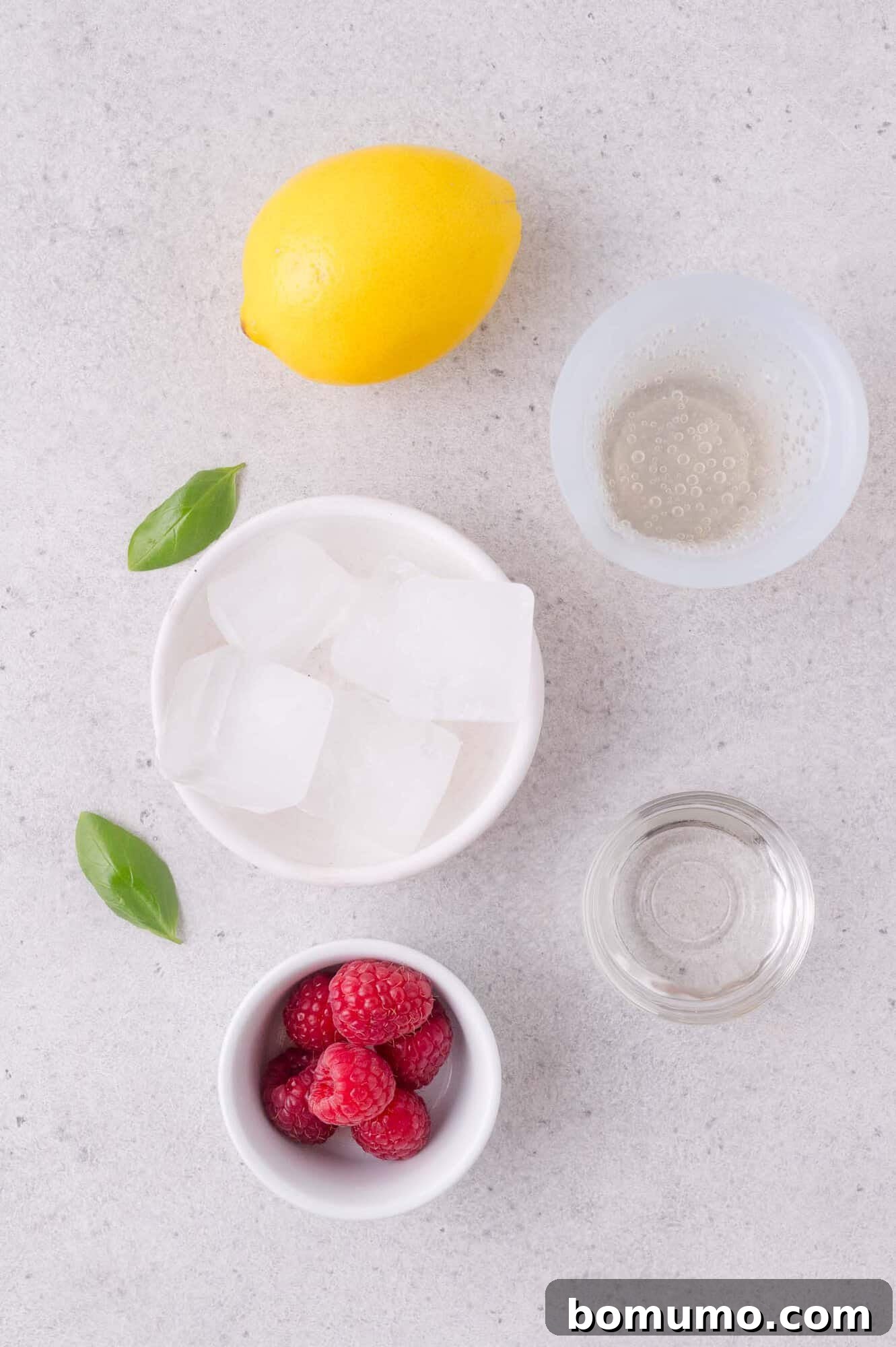 Lemon, glass of club soda, basil leaves, bowl of ice cubes, bowl of raspberries, and a small glass of clear liquid on a light surface.
