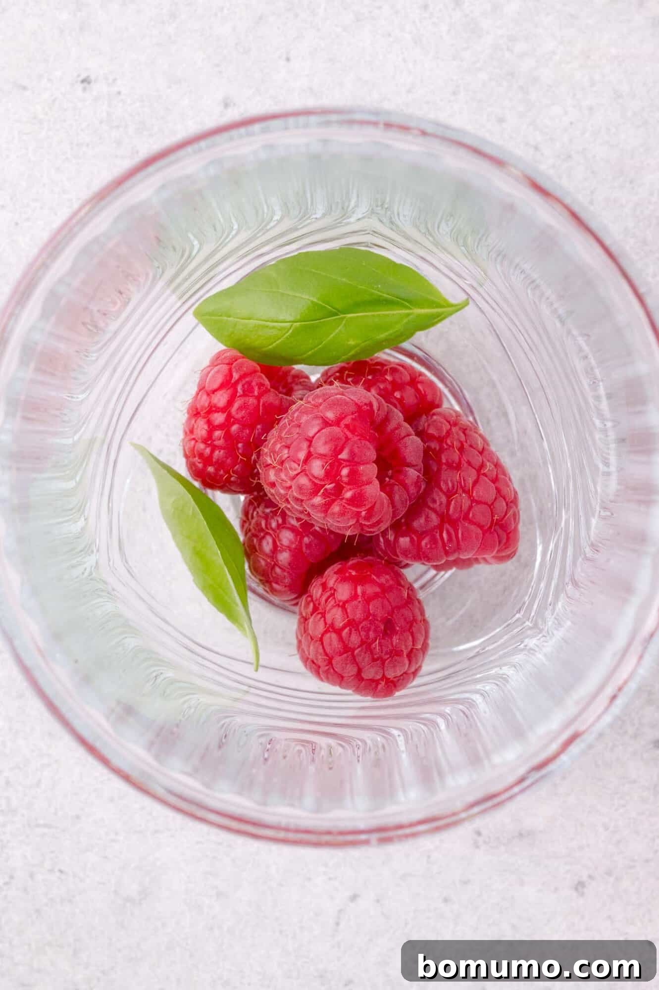 Five fresh raspberries and two green basil leaves in the bottom of a clear glass, viewed from above on a light surface.
