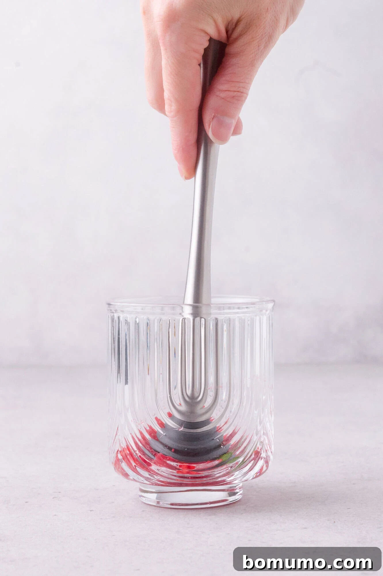 A hand uses a metal muddler to mash red berries at the bottom of a clear, textured glass on a gray surface.