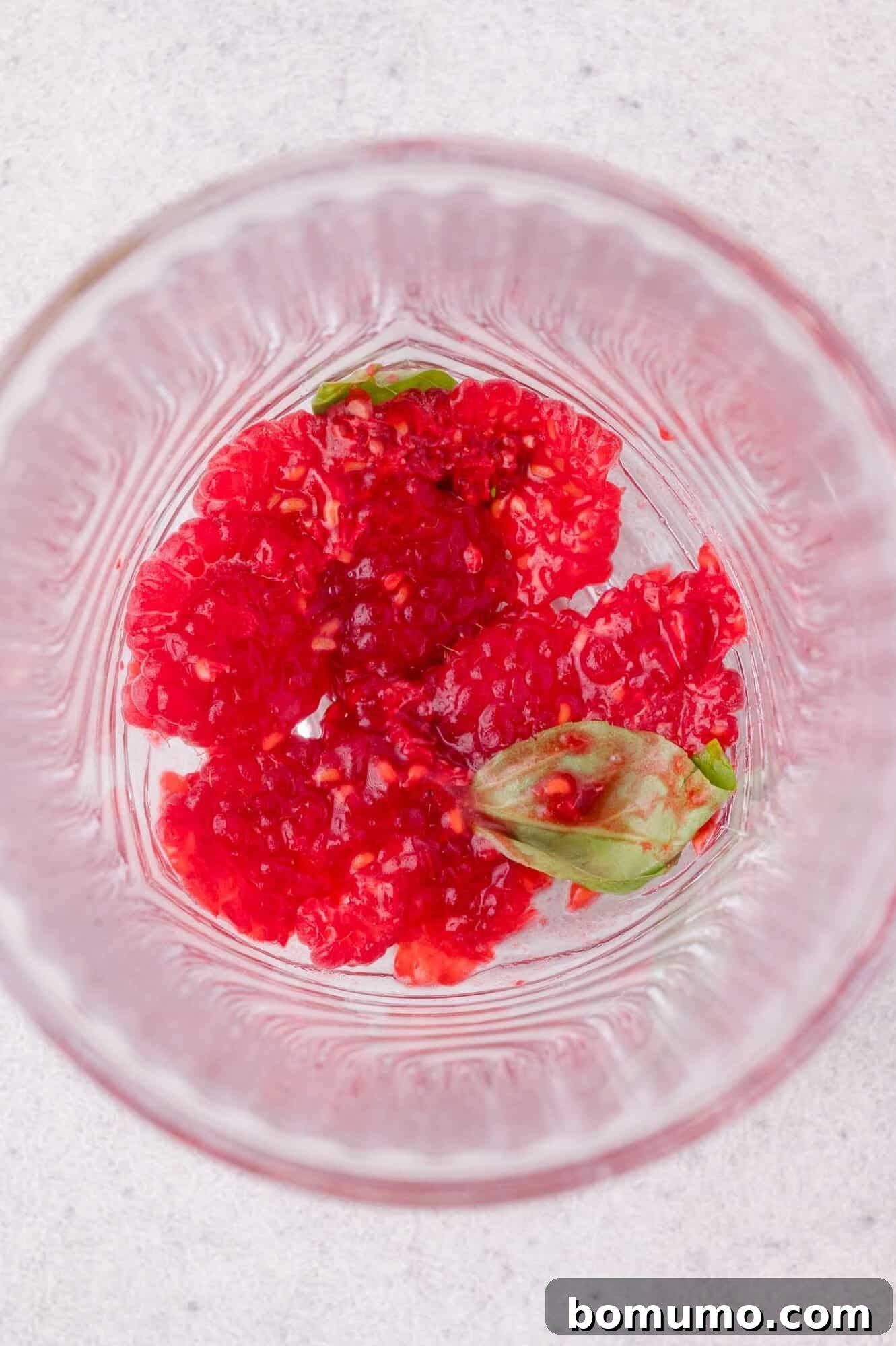 Crushed raspberries and a few basil leaves at the bottom of a clear glass on a light-colored surface.