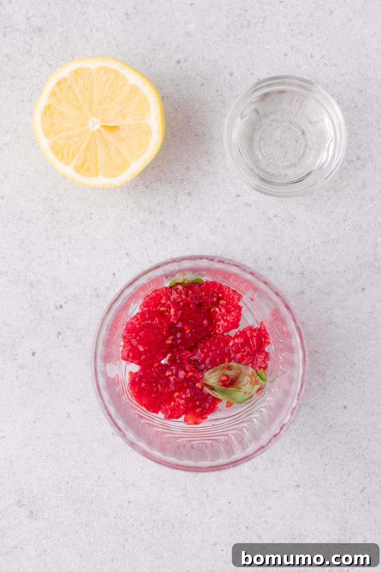 A glass with mashed raspberries and basil, a small bowl of clear liquid, and half a lemon on a light gray surface.