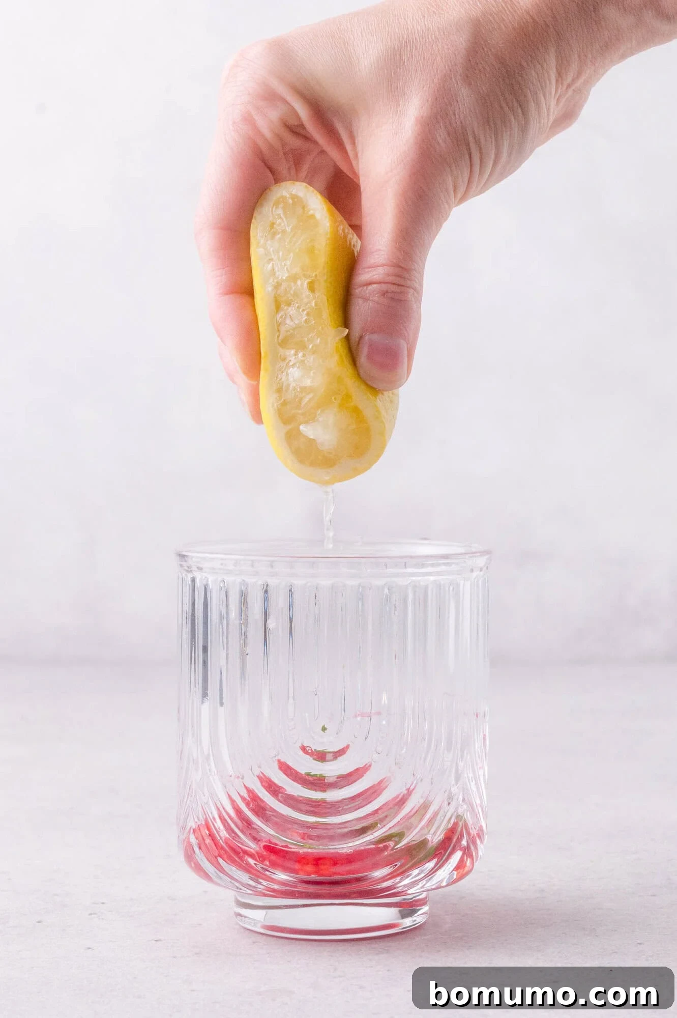 A hand squeezes half a lemon over a clear glass with a red liquid at the bottom, against a light background.