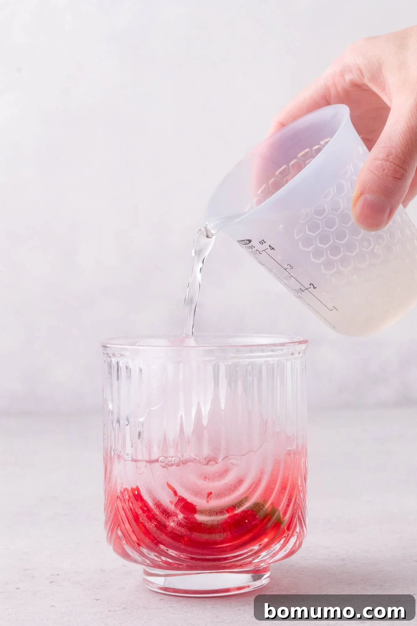 A hand pours sparkling water from a measuring cup into a glass containing red syrup and ice on a light surface.