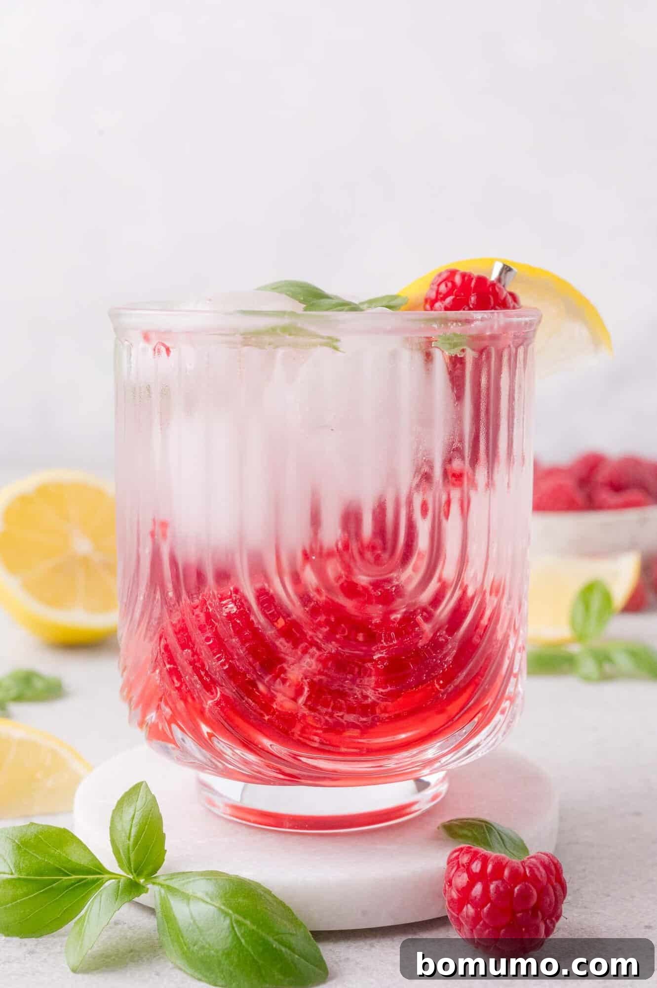 A glass of iced raspberry mocktail garnished with a lemon slice, fresh raspberries, and basil, sits on a white surface with lemon halves and basil leaves around it.