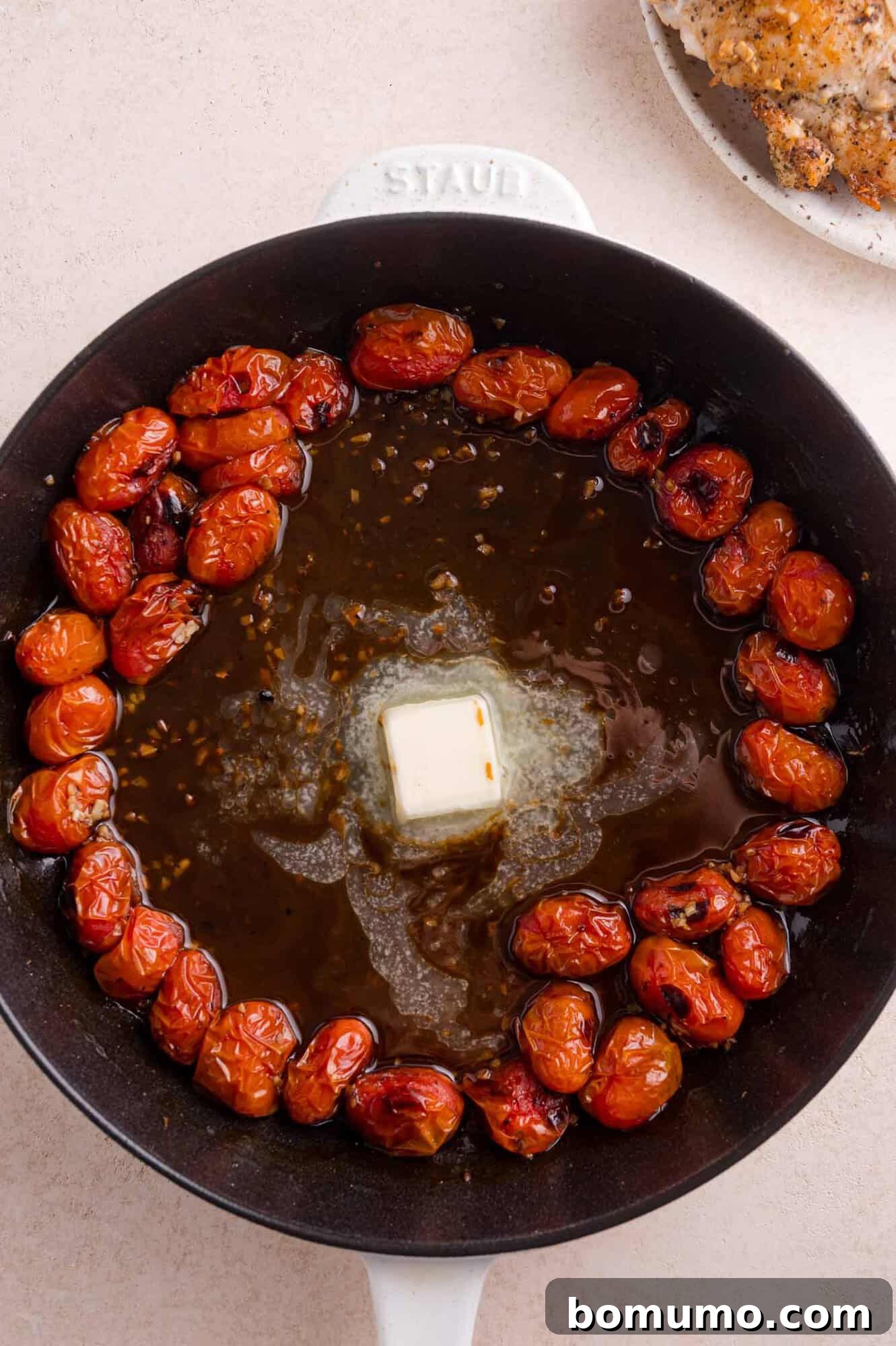 A cast iron pan with roasted cherry tomatoes, melted butter, and sauce in the center, on a beige surface.