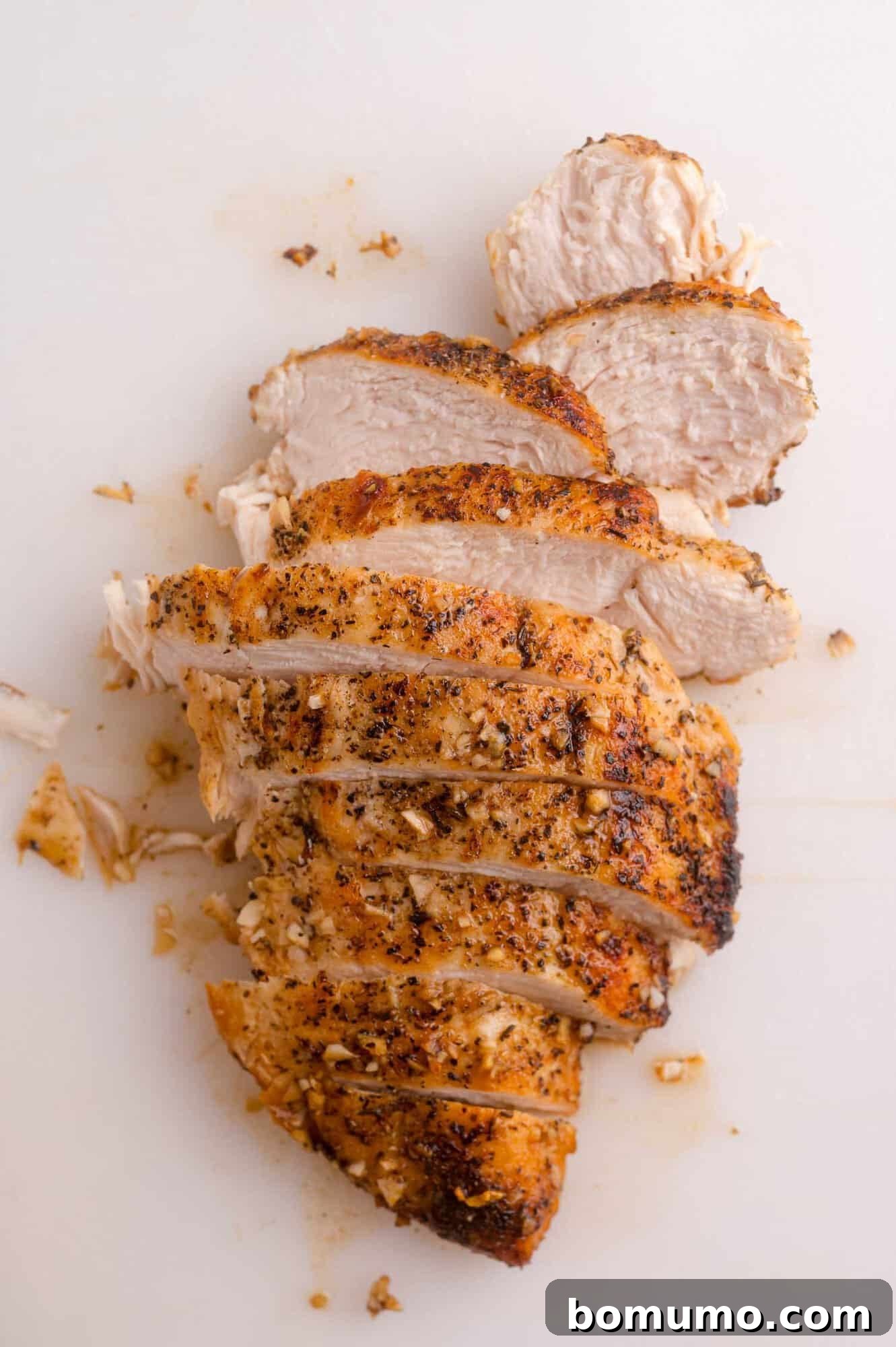 Sliced, cooked chicken breast seasoned with herbs and spices, arranged on a white surface.