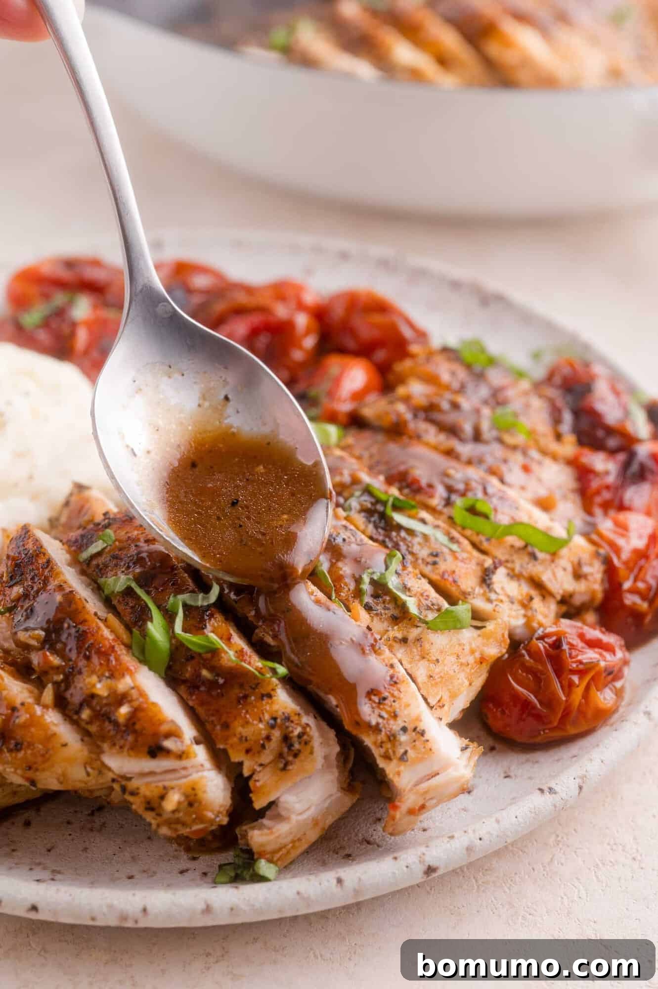 A spoon drizzles sauce over sliced grilled chicken breast, garnished with chopped herbs, with roasted tomatoes and a serving of mashed potatoes on the plate.