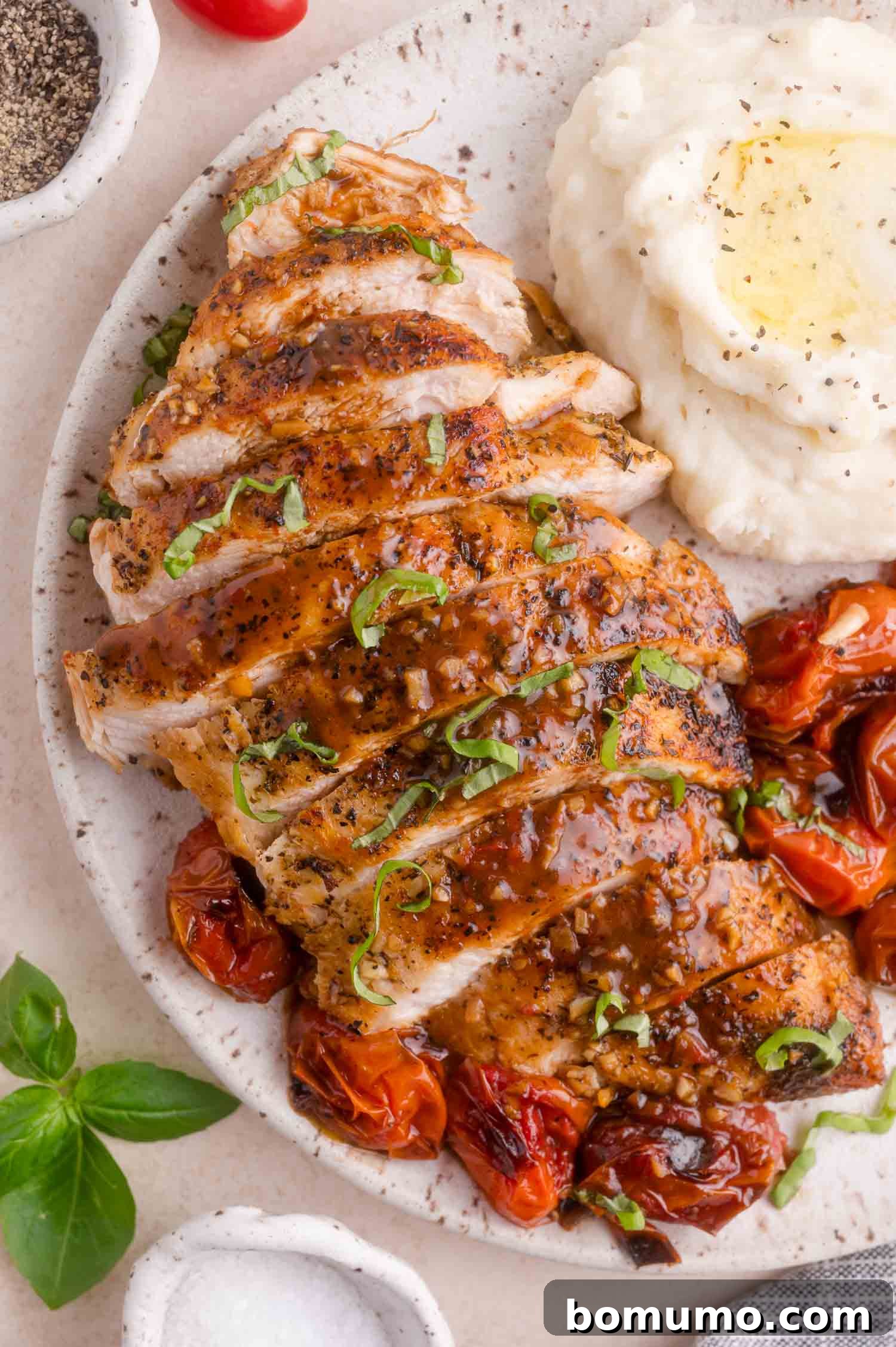 Sliced seasoned chicken breast with sauce, roasted tomatoes, and a serving of mashed potatoes on a white plate.