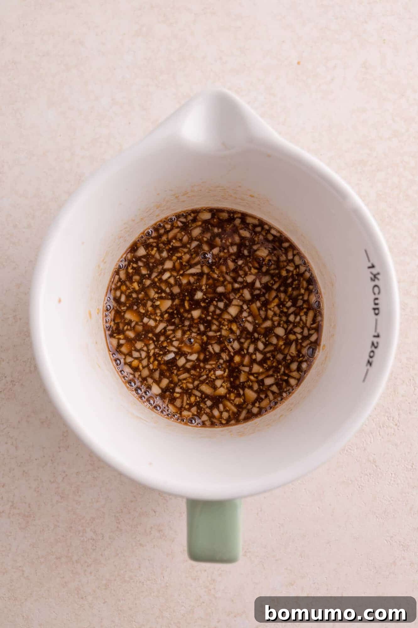 A white measuring cup containing a mixture of finely chopped garlic and balsamic-based sauce, viewed from above on a light surface.