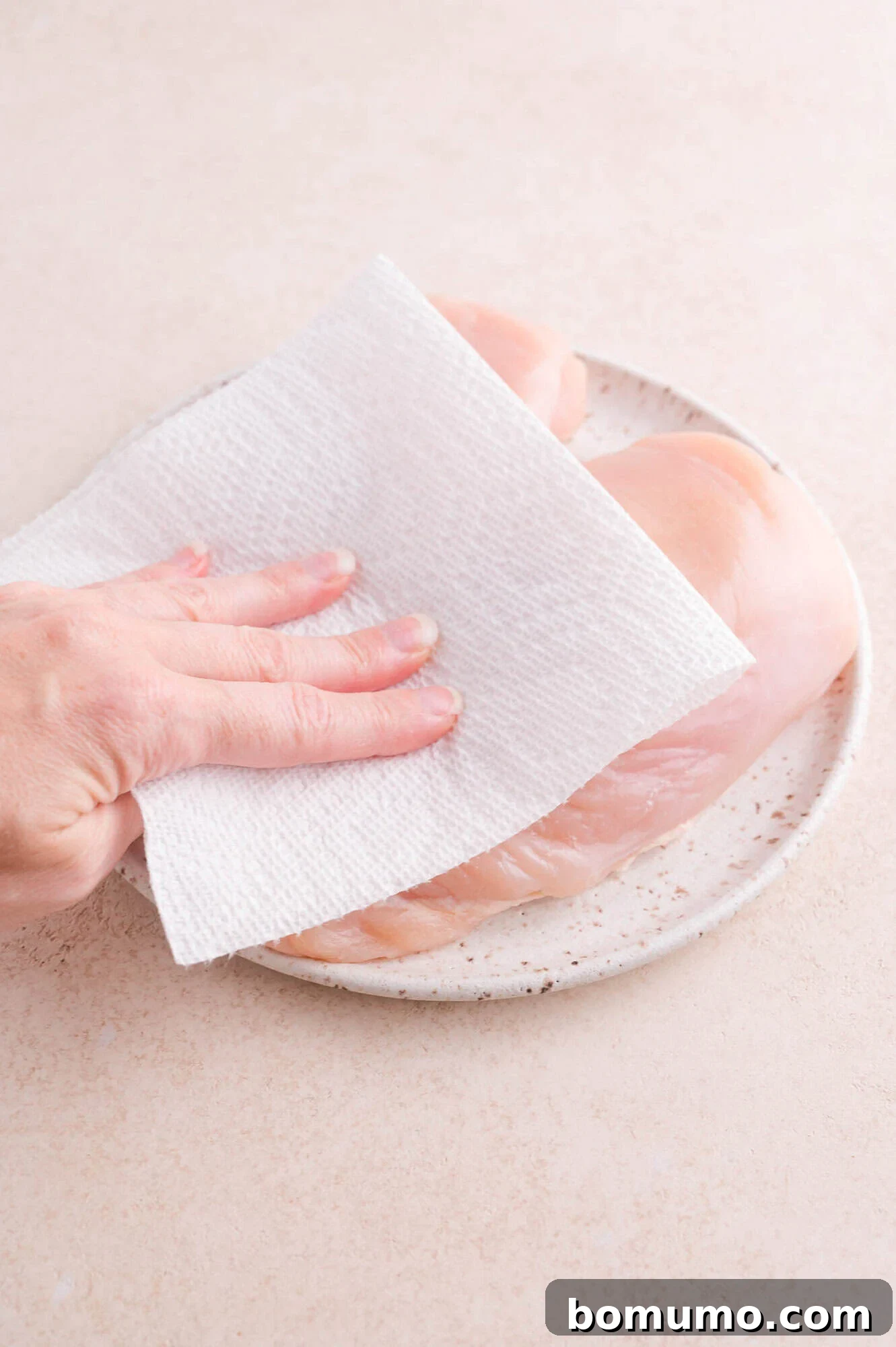 A hand uses a paper towel to pat raw chicken breasts dry on a speckled plate.