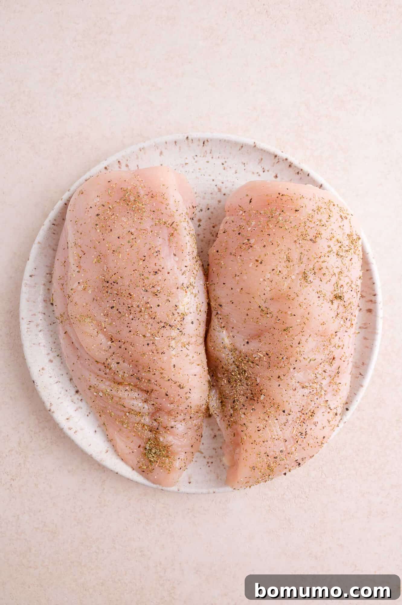 Two raw chicken breasts seasoned with pepper and herbs on a white plate, placed on a light-colored surface.