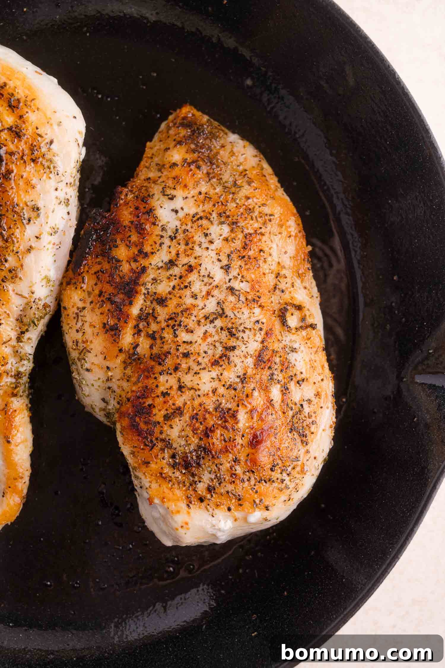 A seasoned, cooked chicken breast sits in a black cast iron skillet, showing a golden-brown, seared surface.