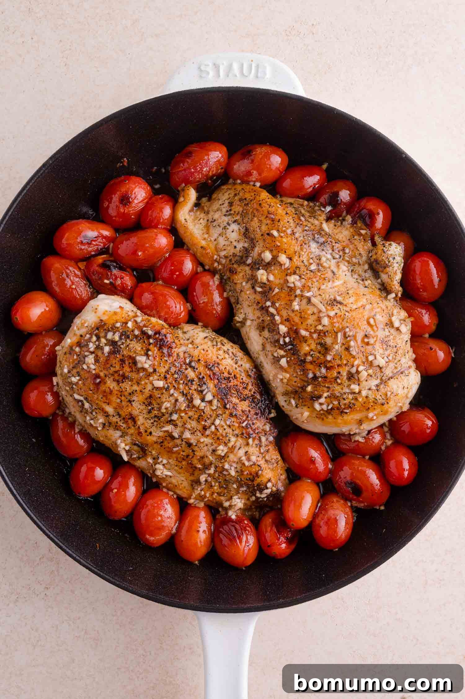 Two seasoned chicken breasts and roasted cherry tomatoes in a black skillet with a white handle.