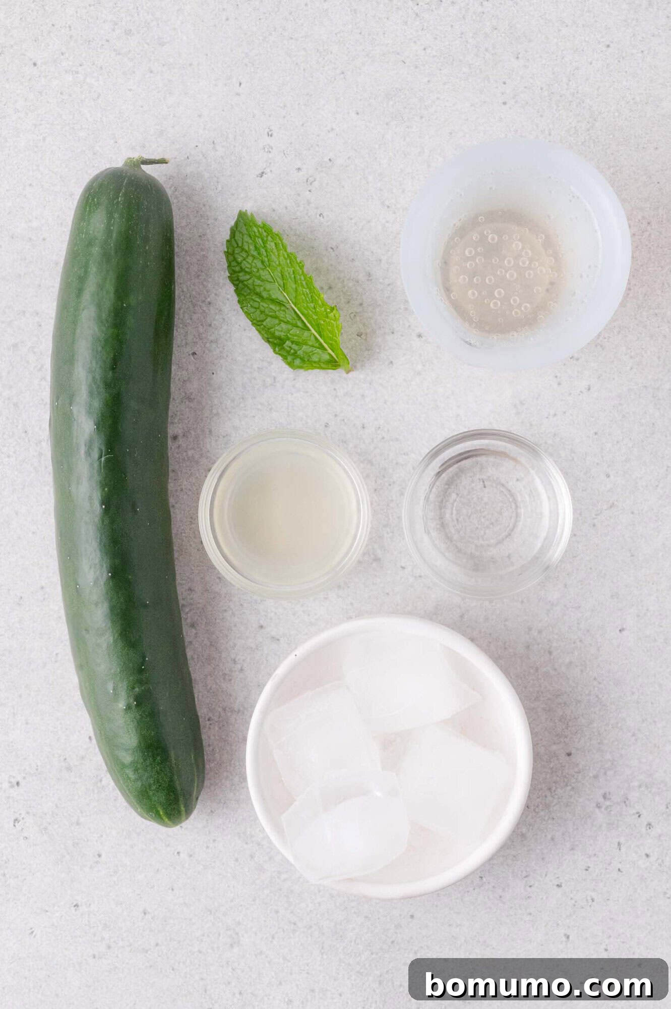 Cucumber, mint leaf, ice cubes in a bowl, and three small bowls containing clear liquids arranged on a light gray surface.