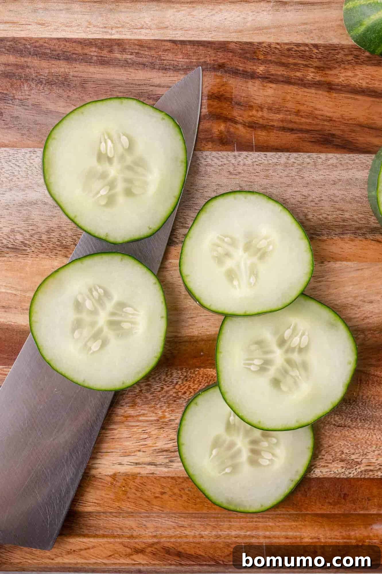 Five cucumber slices and a kitchen knife are on a wooden cutting board.