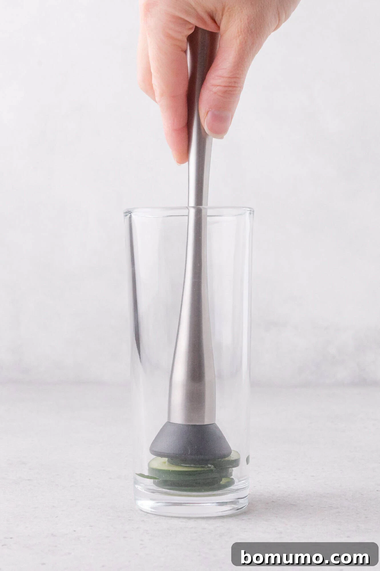 A hand uses a metal muddler to crush lime slices at the bottom of a clear glass on a light surface.