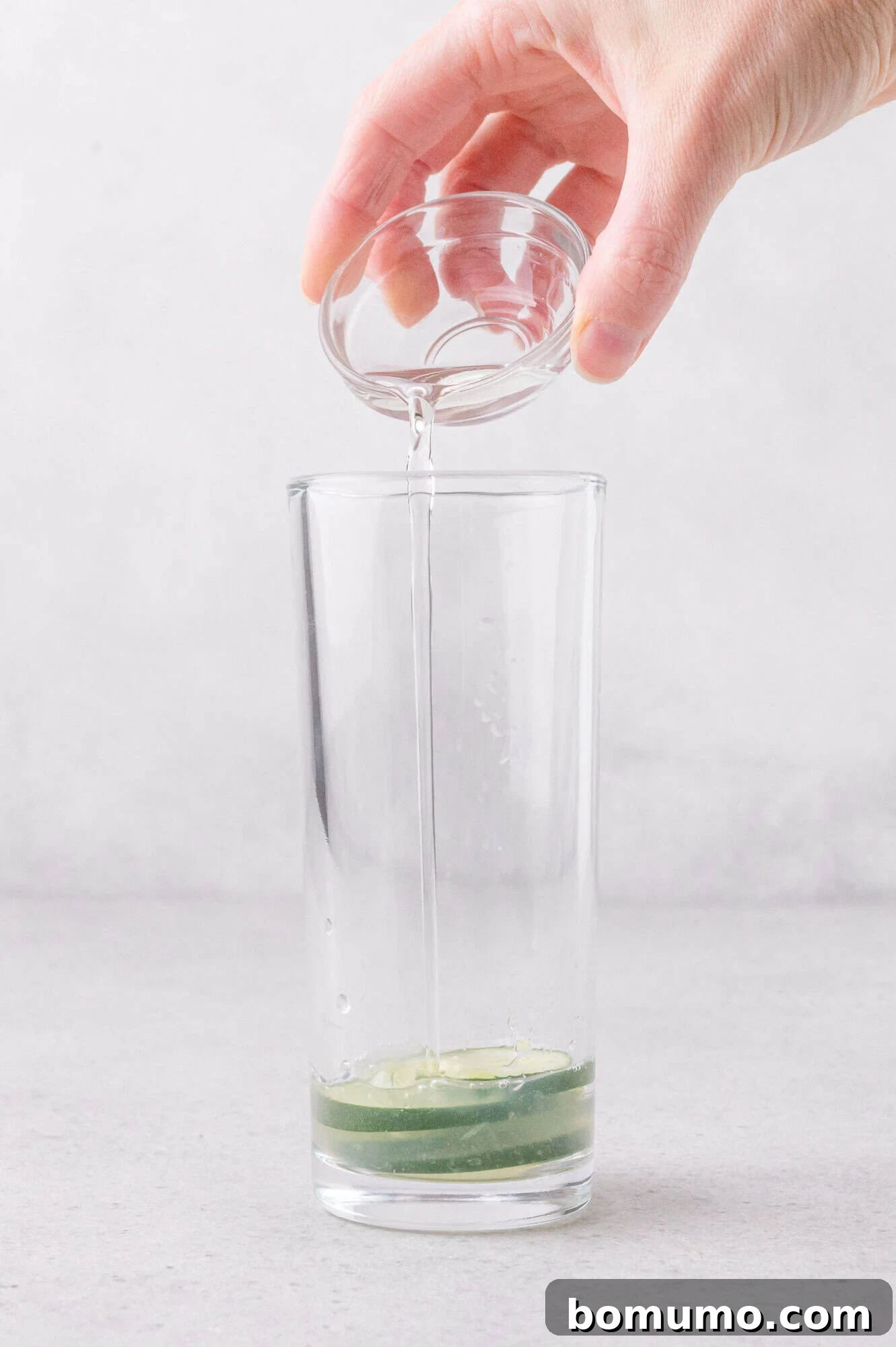A hand pours clear liquid from a small glass bowl into a tall glass containing cucumber slices and juice.