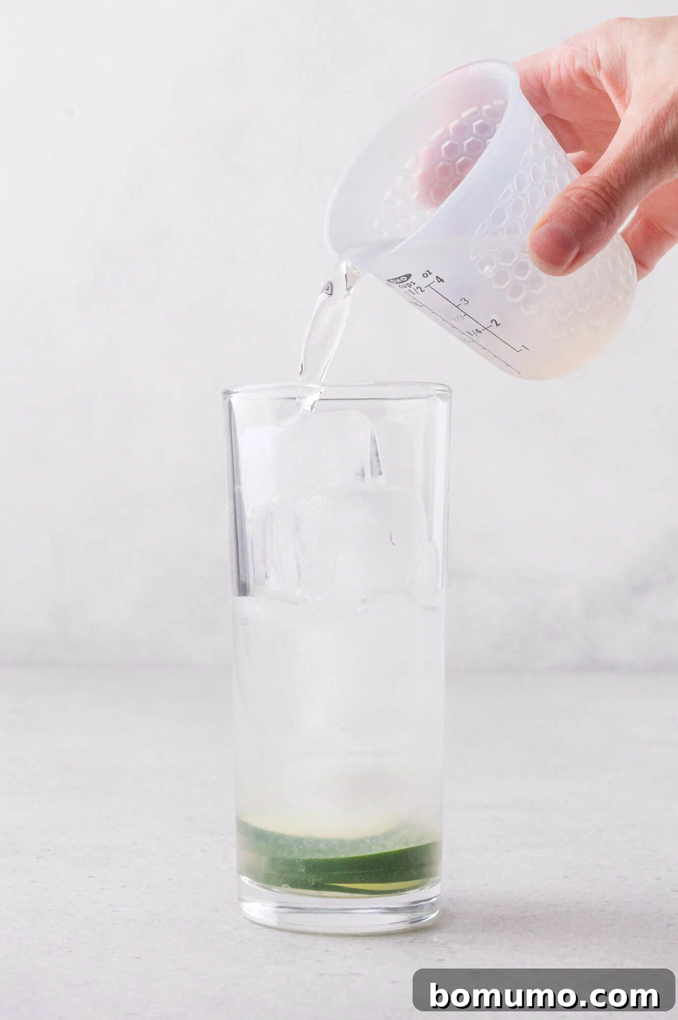 A hand pours clear liquid from a measuring cup into a tall glass filled with ice and a small amount of cucumbers at the bottom.