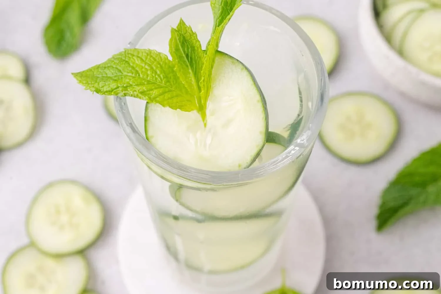 A glass of cucumber lime cooler with cucumber slices and a sprig of mint, placed on a white coaster, surrounded by cucumber slices and mint leaves.