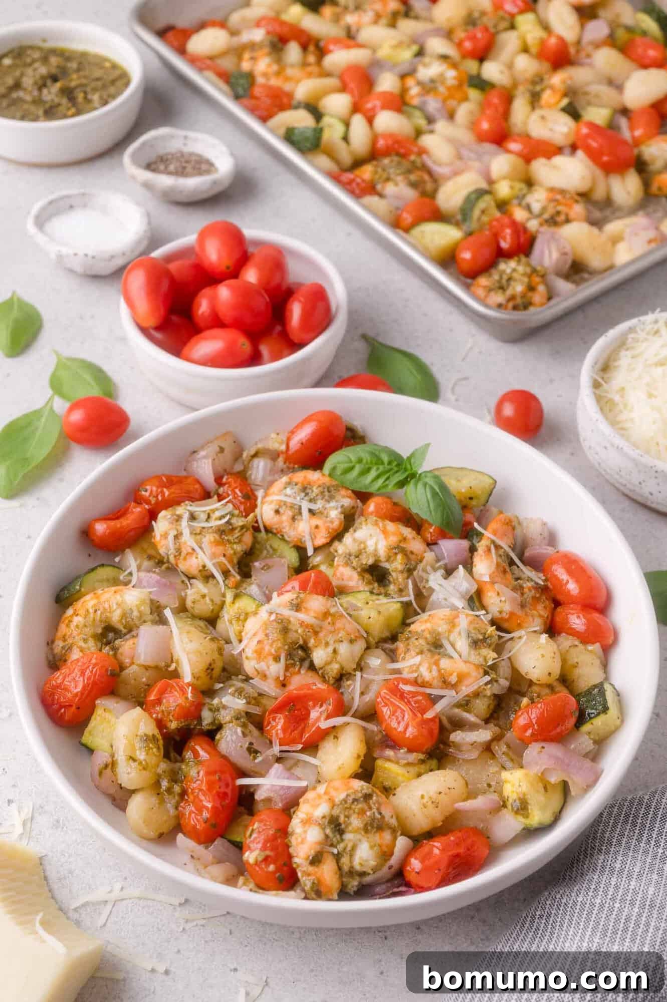 Bowl of gnocchi with shrimp, cherry tomatoes, zucchini, red onion, and herbs, with a tray of the same dish, grated cheese, and fresh tomatoes in the background.