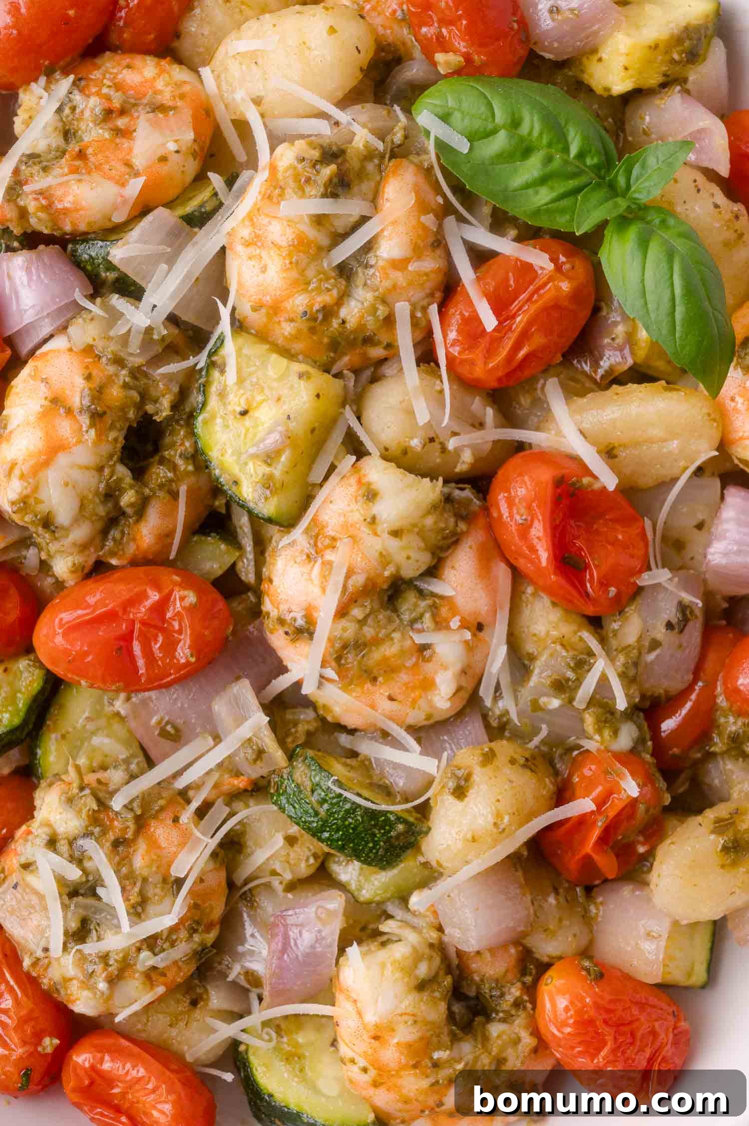Cooked shrimp with cherry tomatoes, zucchini, onions, shredded cheese, and pesto, garnished with a basil sprig.