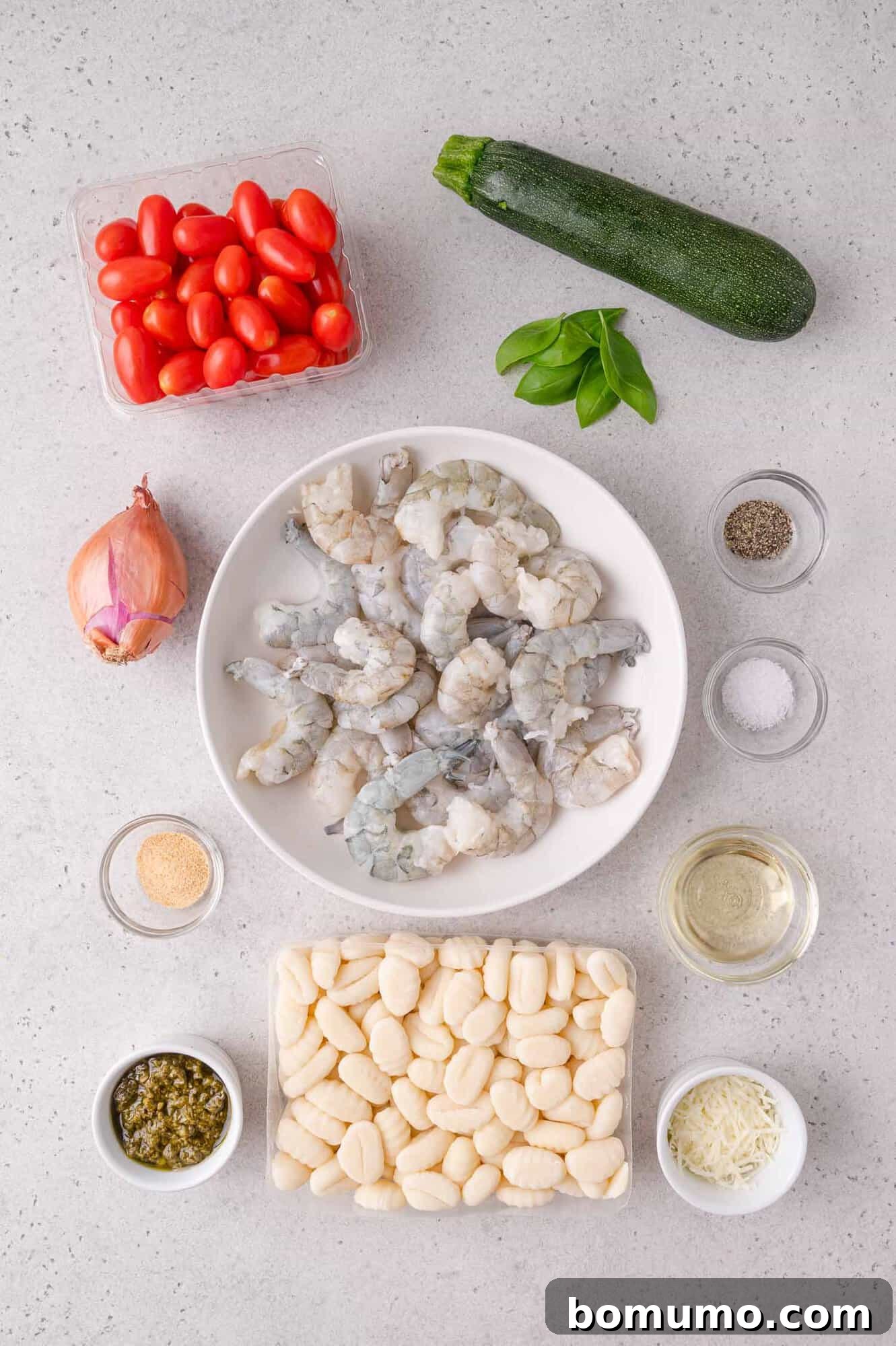 Ingredients for a shrimp and gnocchi dish arranged on a countertop: raw shrimp, gnocchi, cherry tomatoes, zucchini, shallot, basil, pesto, grated cheese, seasoning, and oil.