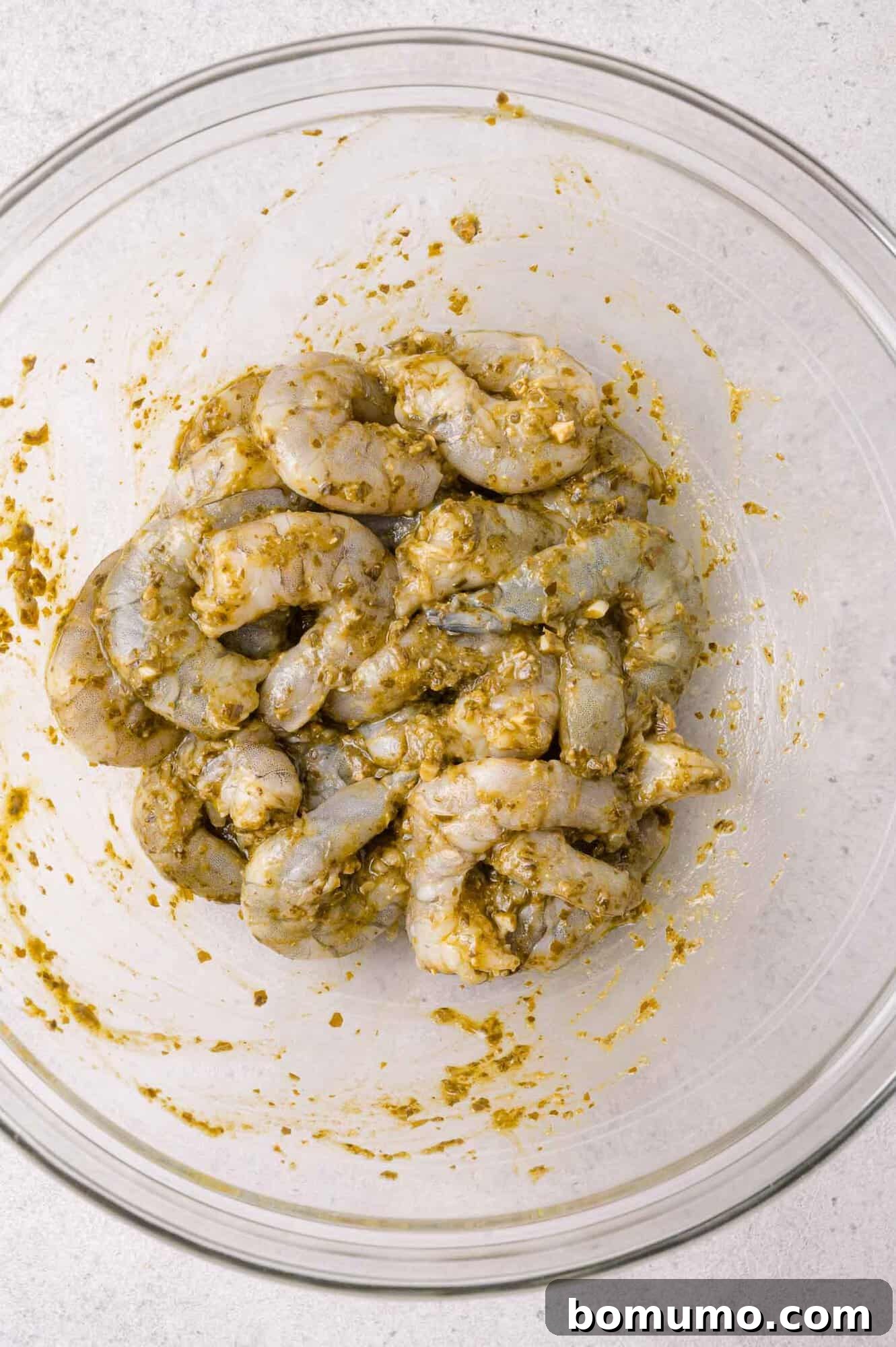 Raw shrimp marinating in pesto in a clear glass bowl on a light-colored surface.