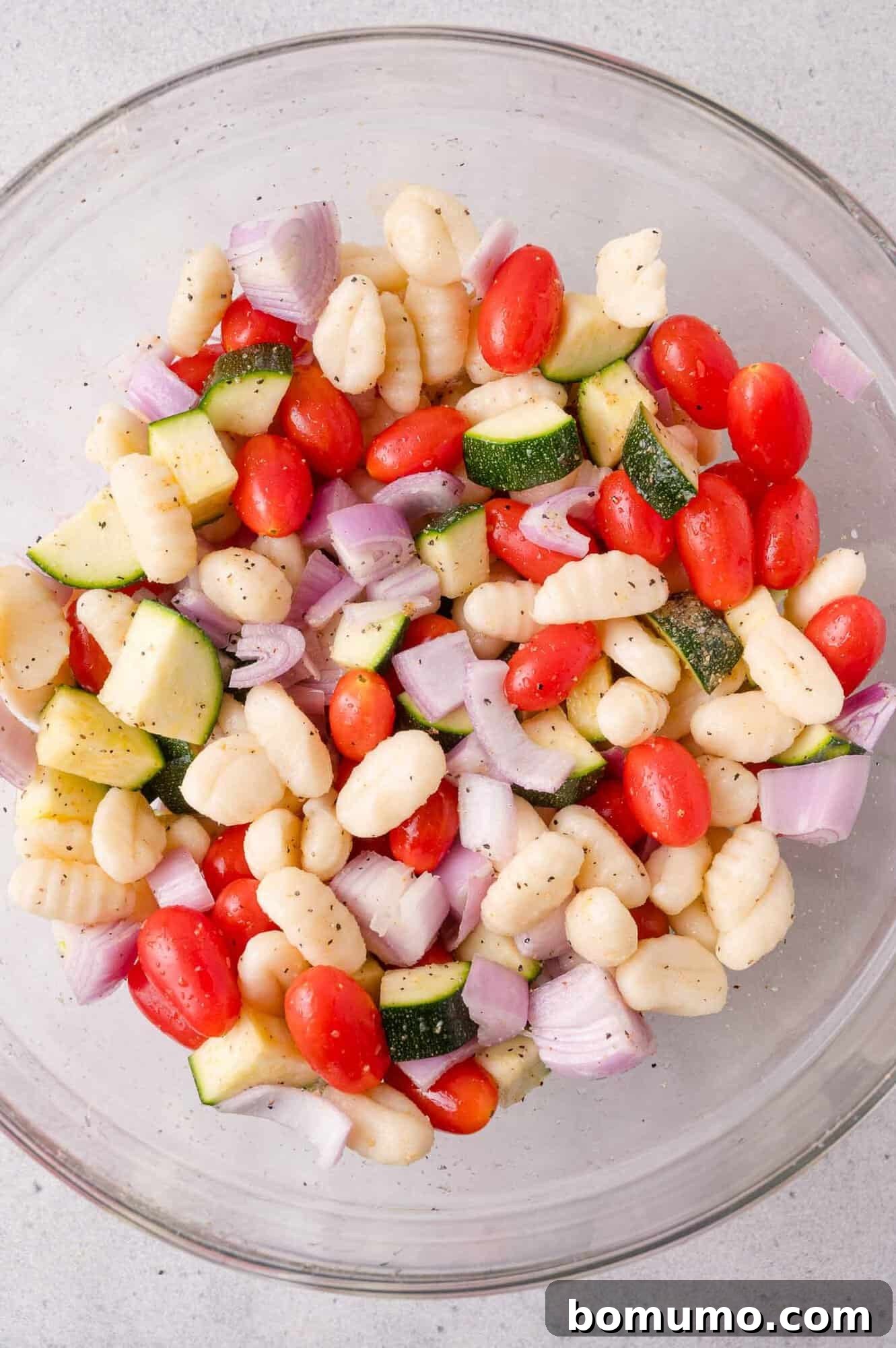 A glass bowl filled with uncooked gnocchi, halved cherry tomatoes, chopped zucchini, and diced red onions, seasoned with black pepper.