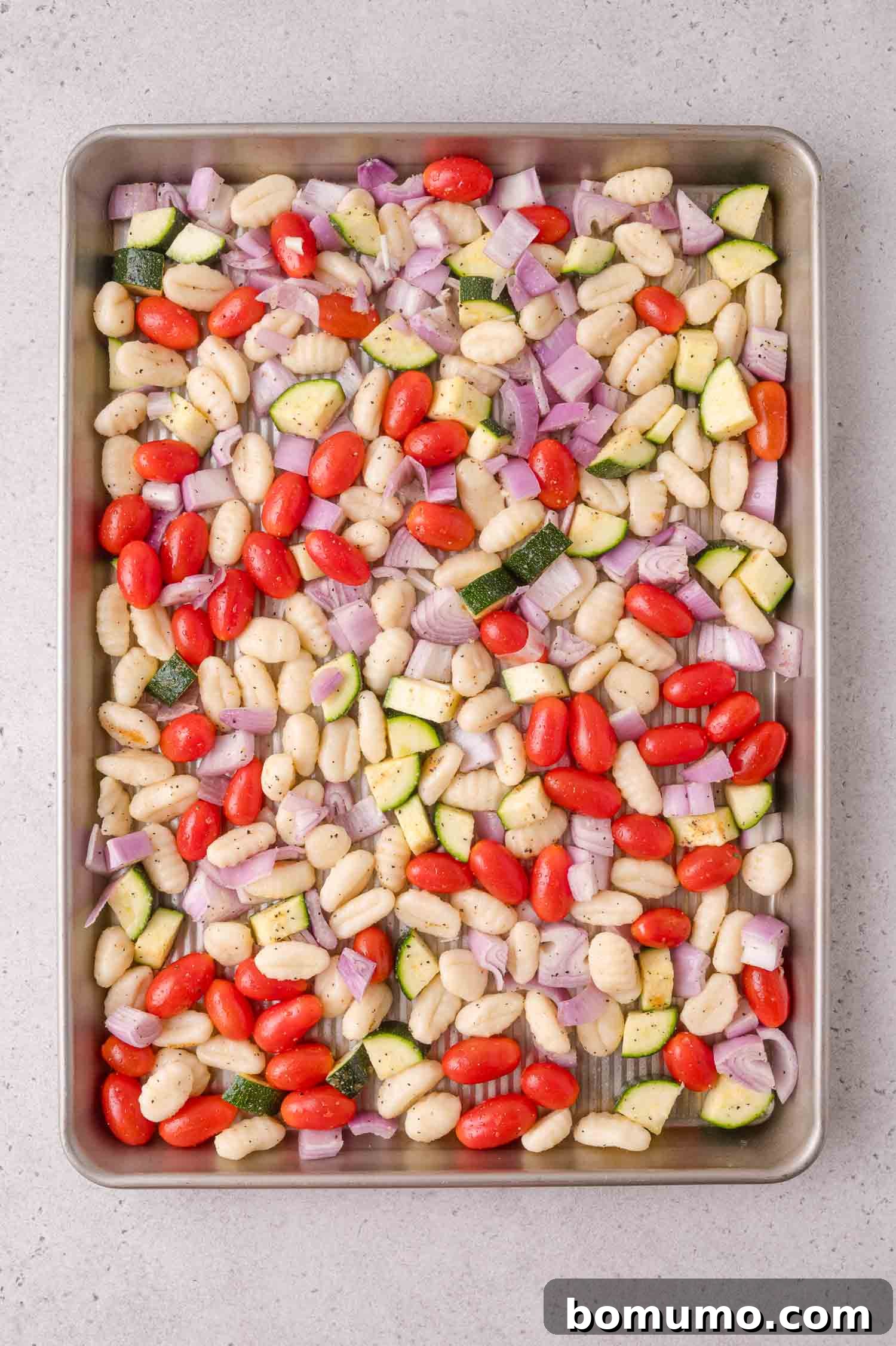 A baking sheet with uncooked gnocchi, cherry tomatoes, diced red onion, and zucchini, seasoned and ready to roast.