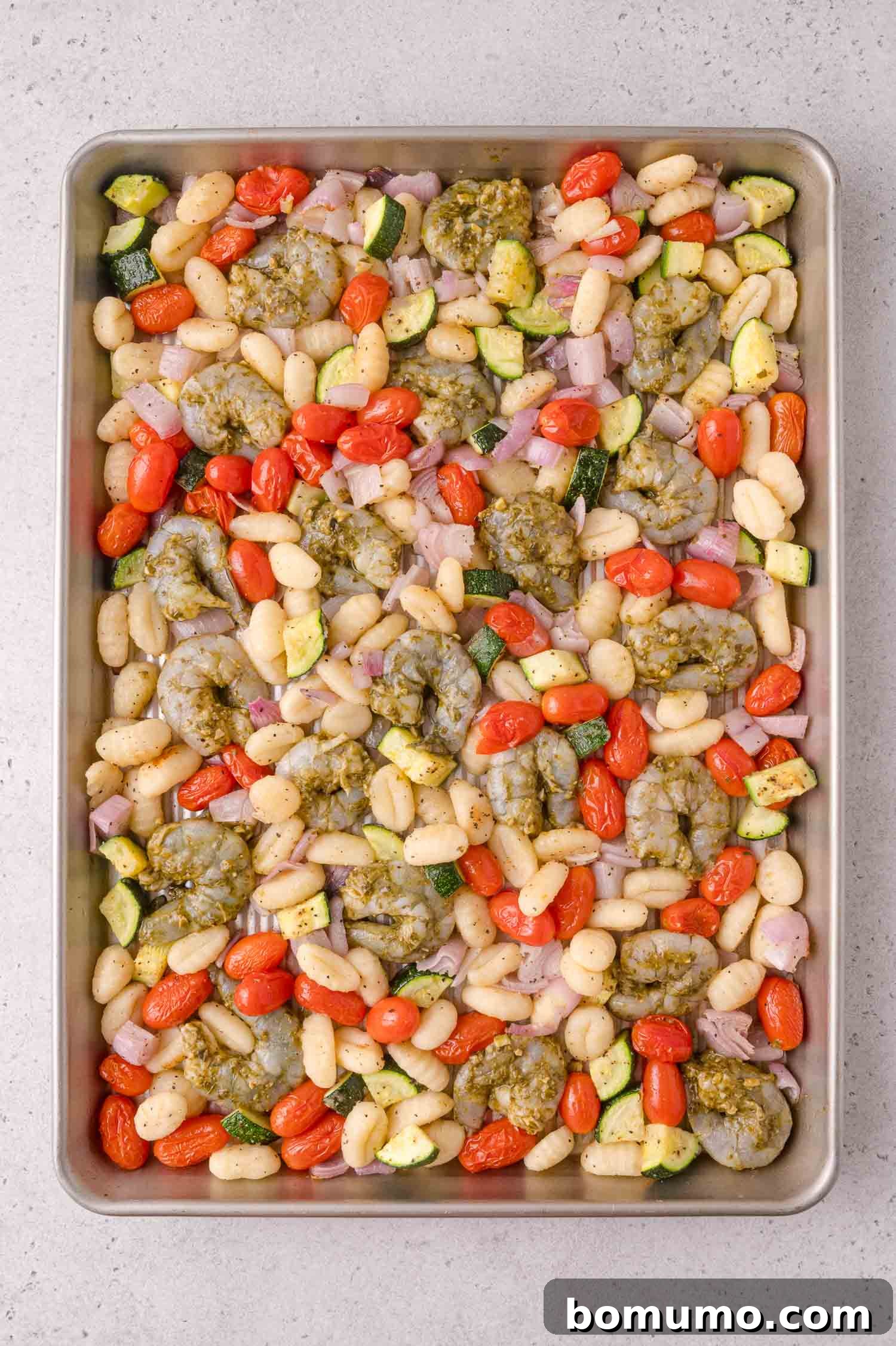 Sheet pan with raw shrimp, gnocchi, cherry tomatoes, zucchini, red onion, and dollops of pesto, ready to be cooked.