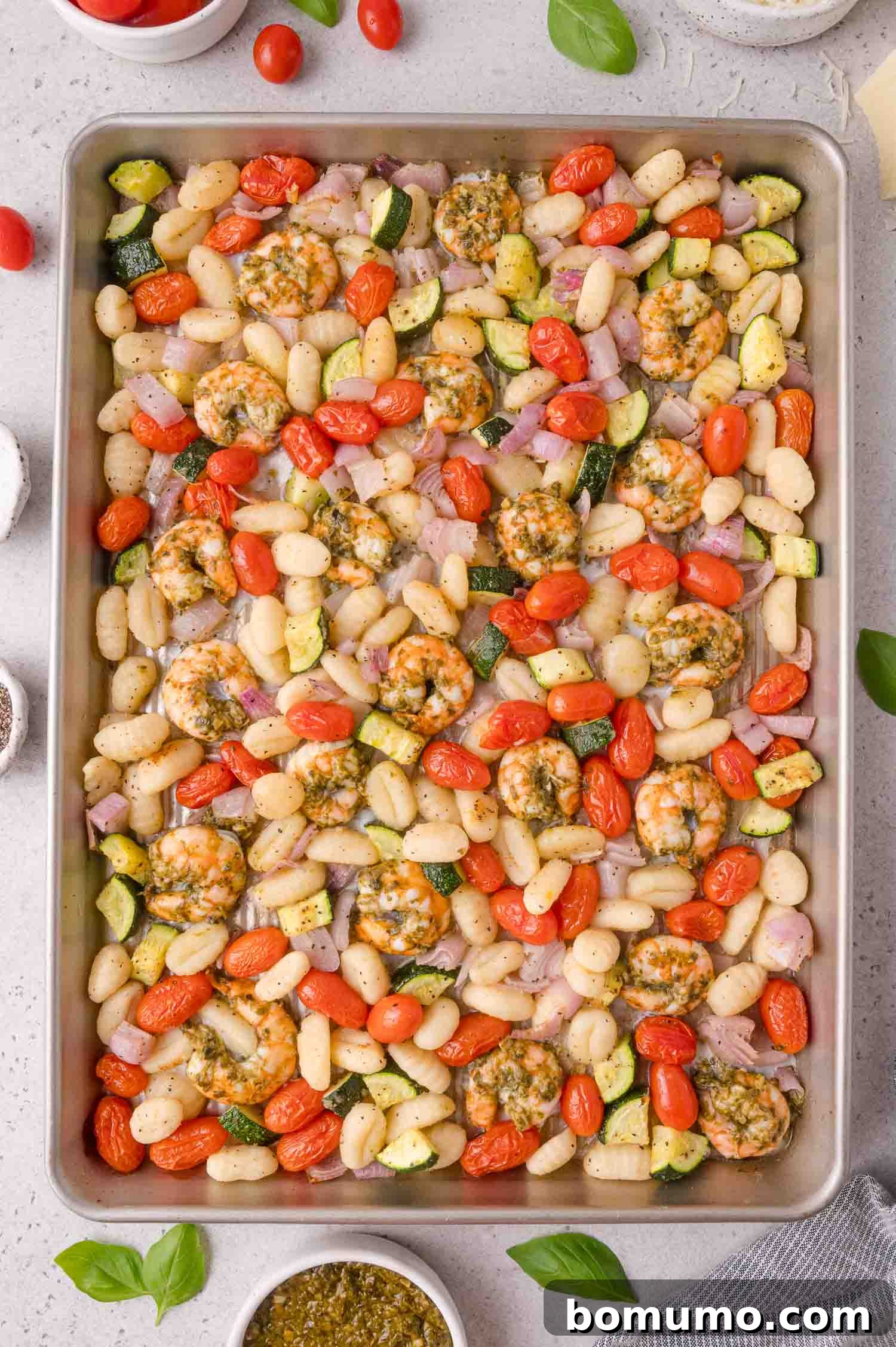 Sheet pan with cooked shrimp, gnocchi, cherry tomatoes, zucchini, and red onion, garnished with herbs. Small bowls of ingredients and basil leaves are visible around the tray.
