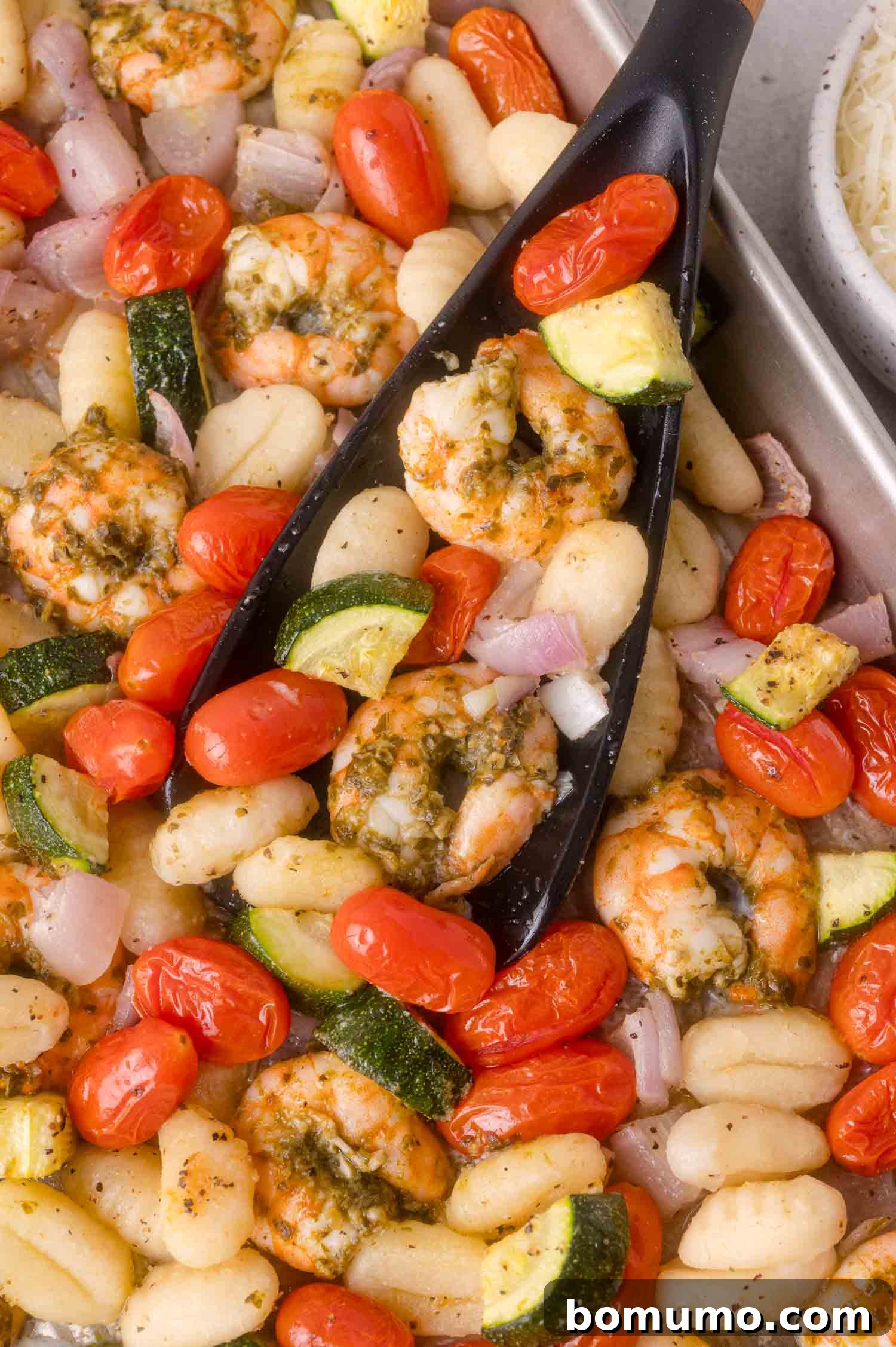 Sheet pan with cooked shrimp, gnocchi, cherry tomatoes, zucchini, and red onions, with a black serving spoon lifting a portion.
