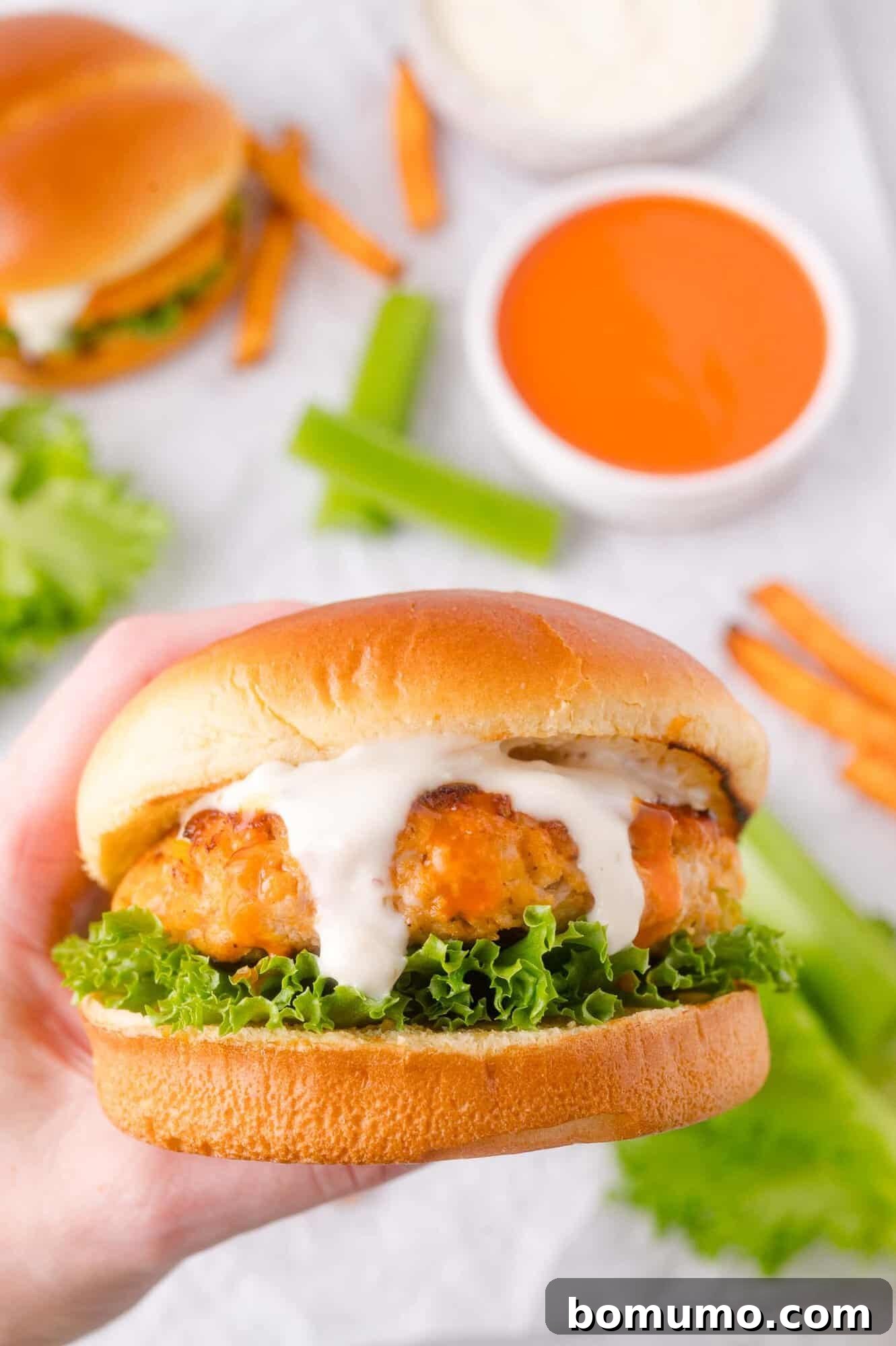 A hand holds a chicken burger with lettuce and ranch sauce, with celery sticks, and dipping sauces in the background.