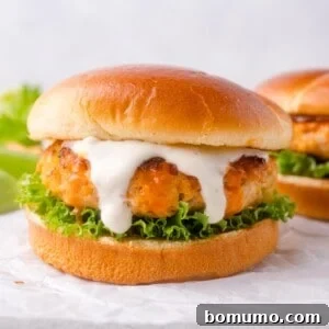 A buffalo chicken burger with lettuce and white sauce is served on a round bun, placed on white parchment paper, with a second burger in the background.