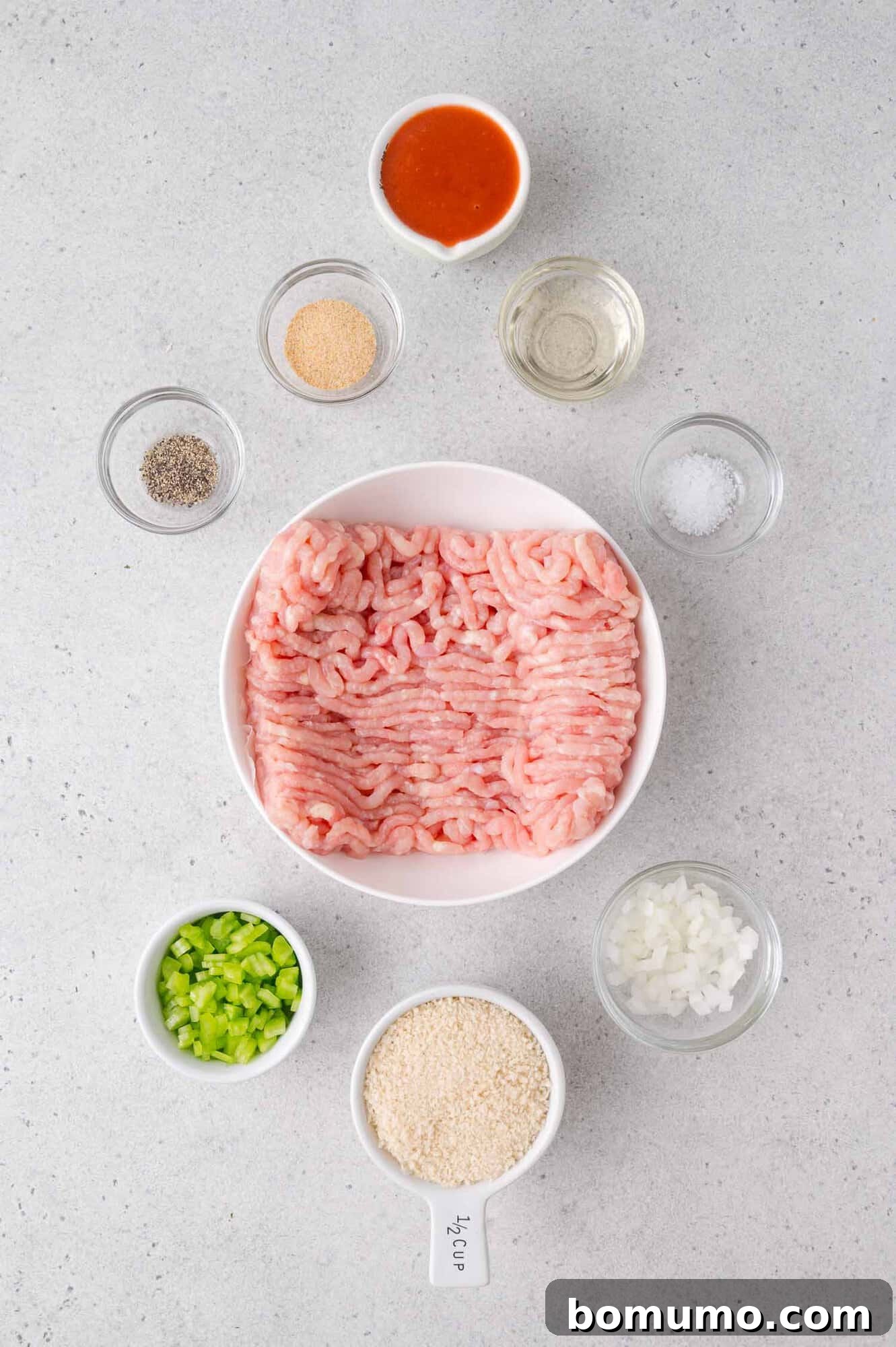 A plate of raw ground chicken is surrounded by small bowls of diced onion, diced celery, breadcrumbs, pepper, garlic powder, salt, hot sauce, and oil on a light countertop.